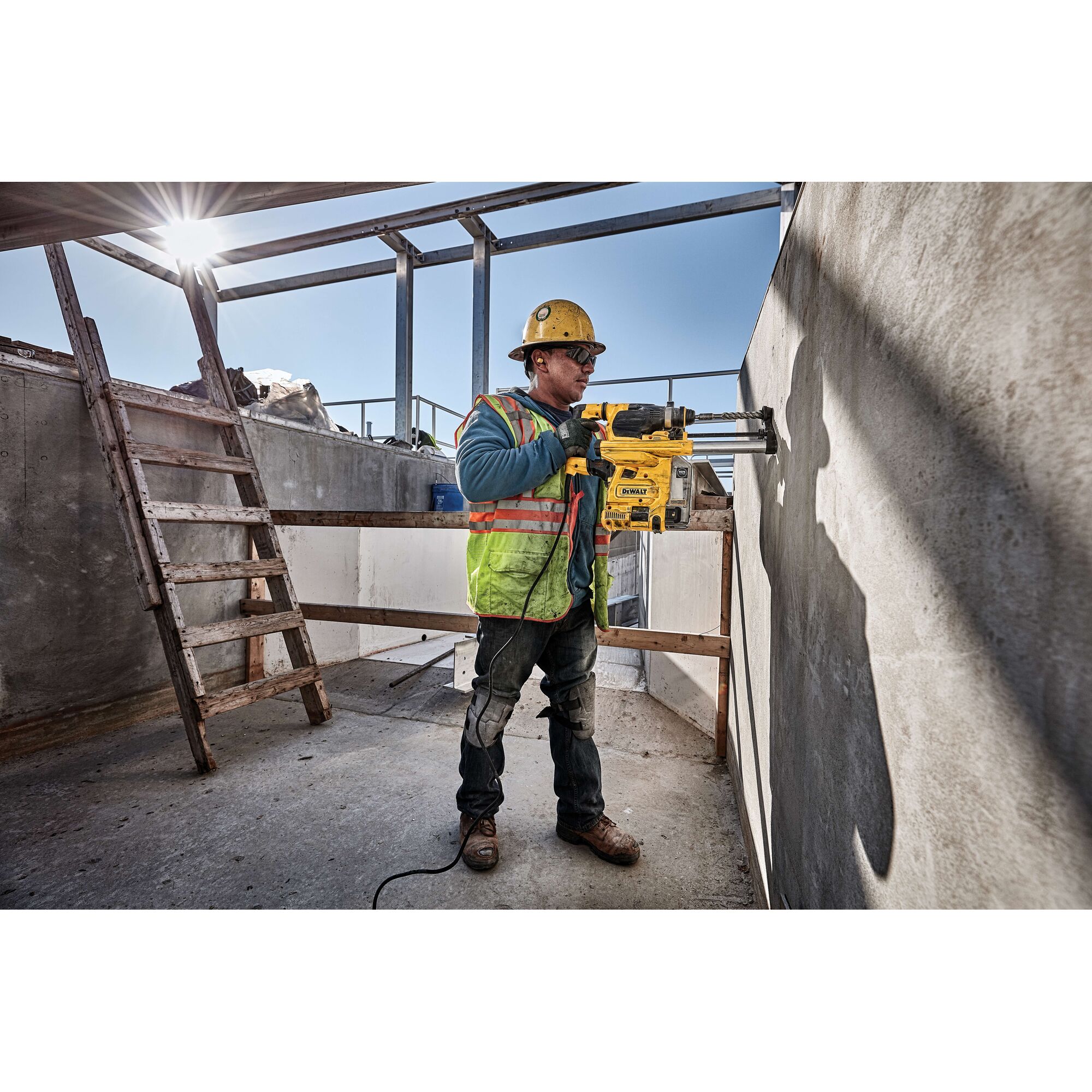 A construction worker wearing a safety helmet and reflective vest uses a DEWALT D25333K power tool to drill into a concrete wall at a construction site.