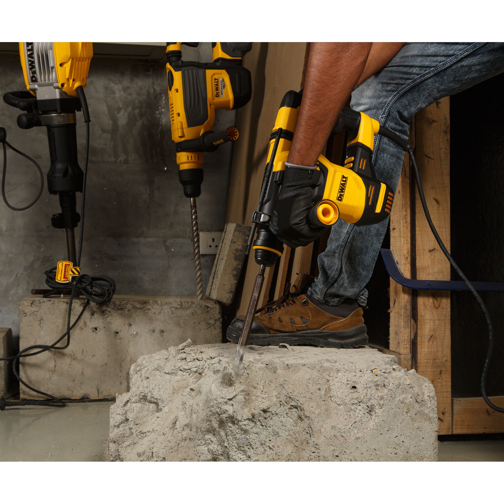 A DEWALT rotary hammer drill D25333K is being used by a person wearing gloves and work boots to drill into a concrete block. Additional DEWALT drills can be seen in the background in a workshop setting.
