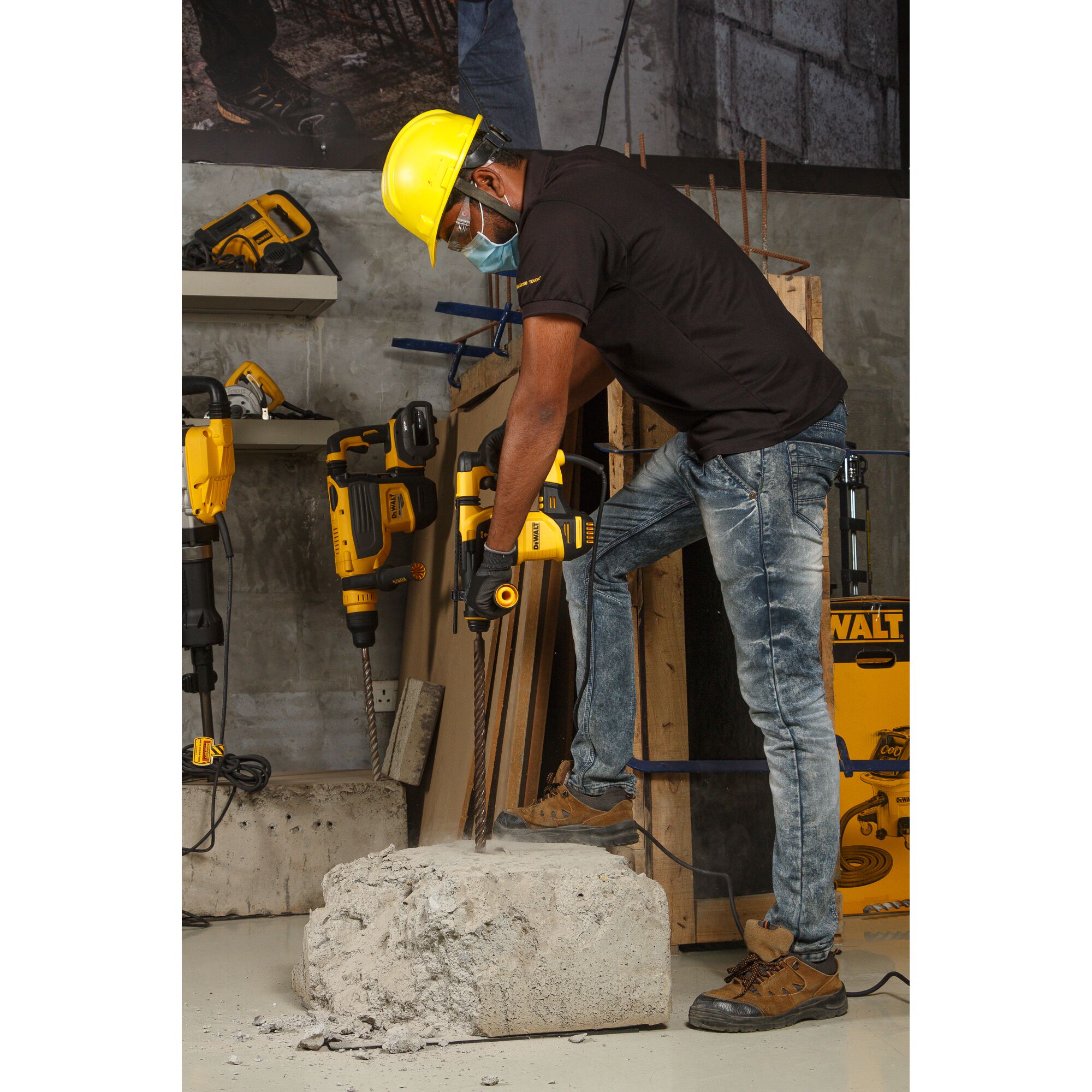 A person wearing a yellow safety helmet and gloves is using a DEWALT power tool to drill into a large concrete block. The individual is standing in a workshop environment with various DEWALT tools and equipment visible in the background.