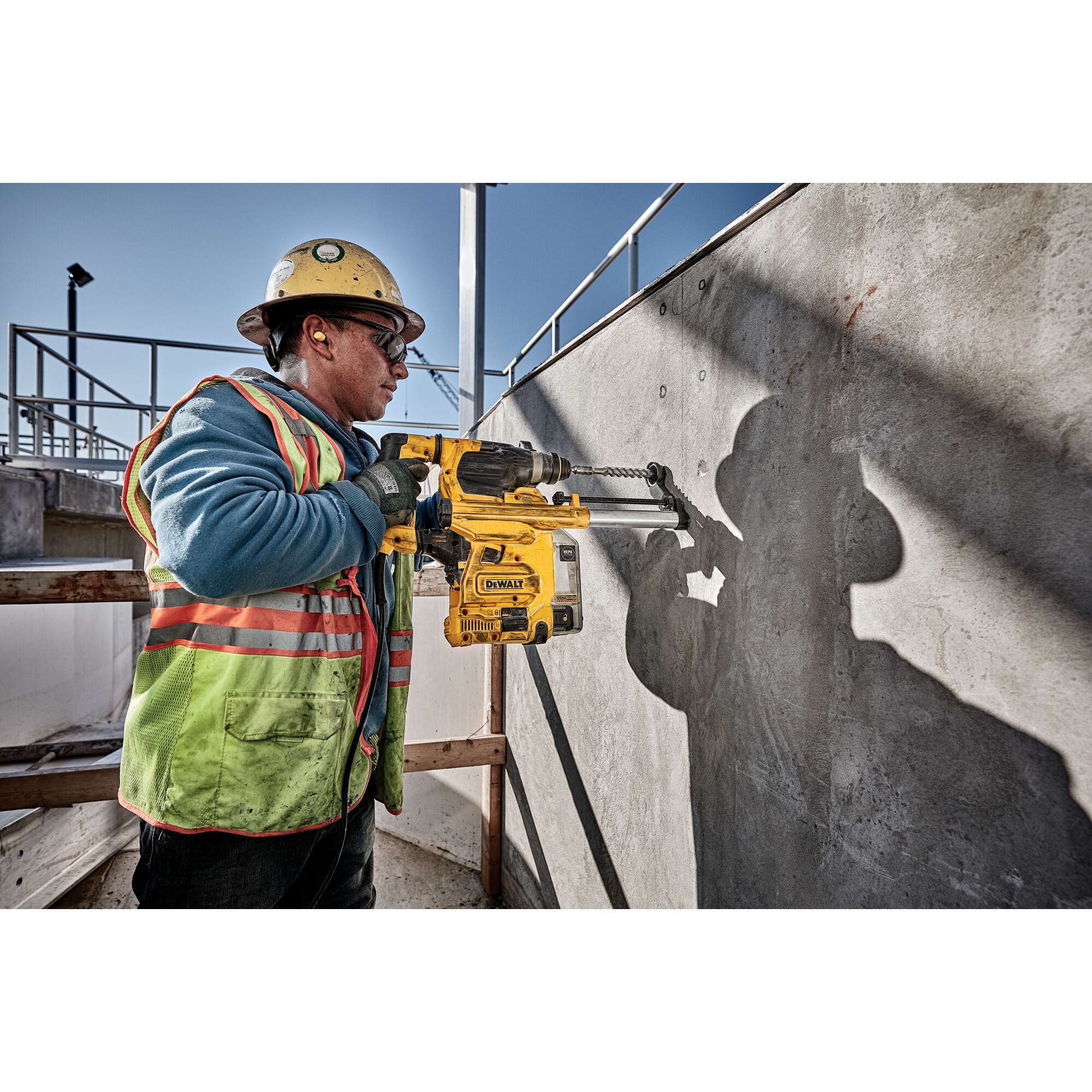 A construction worker wearing a helmet and safety vest is using a DEWALT D25333K rotary hammer drill to bore into a concrete wall at an outdoor construction site.