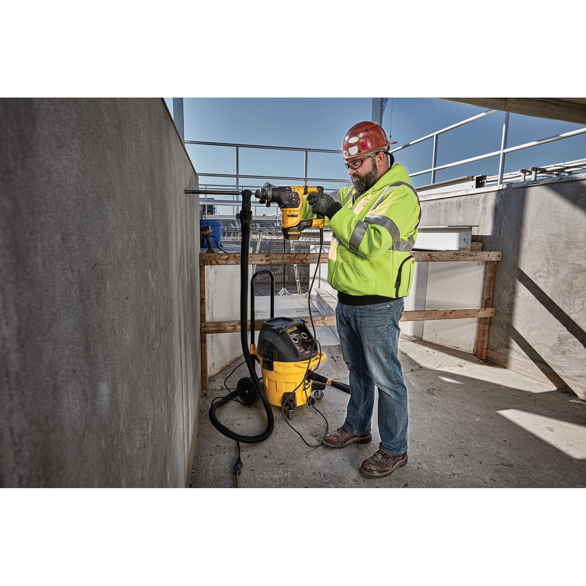 A person wearing a high-visibility jacket and a red hard hat is using a DEWALT power tool to drill into a concrete wall at a construction site. A yellow and black DEWALT vacuum is connected to the tool to manage dust.
