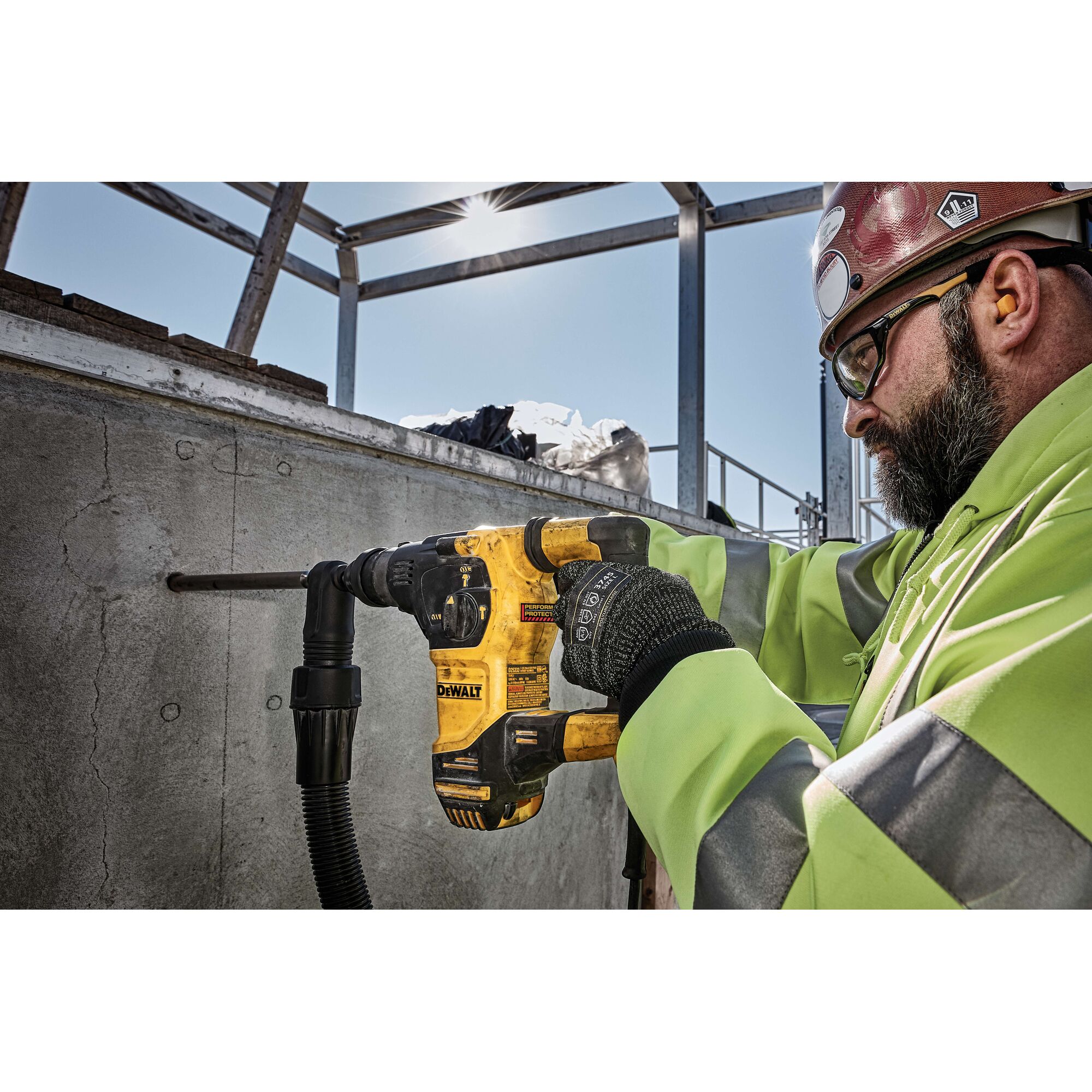 A person wearing a high-visibility safety jacket and gloves is using a yellow DEWALT rotary hammer drill to bore a hole into a concrete wall at a construction site. The person's face is blurred for privacy.