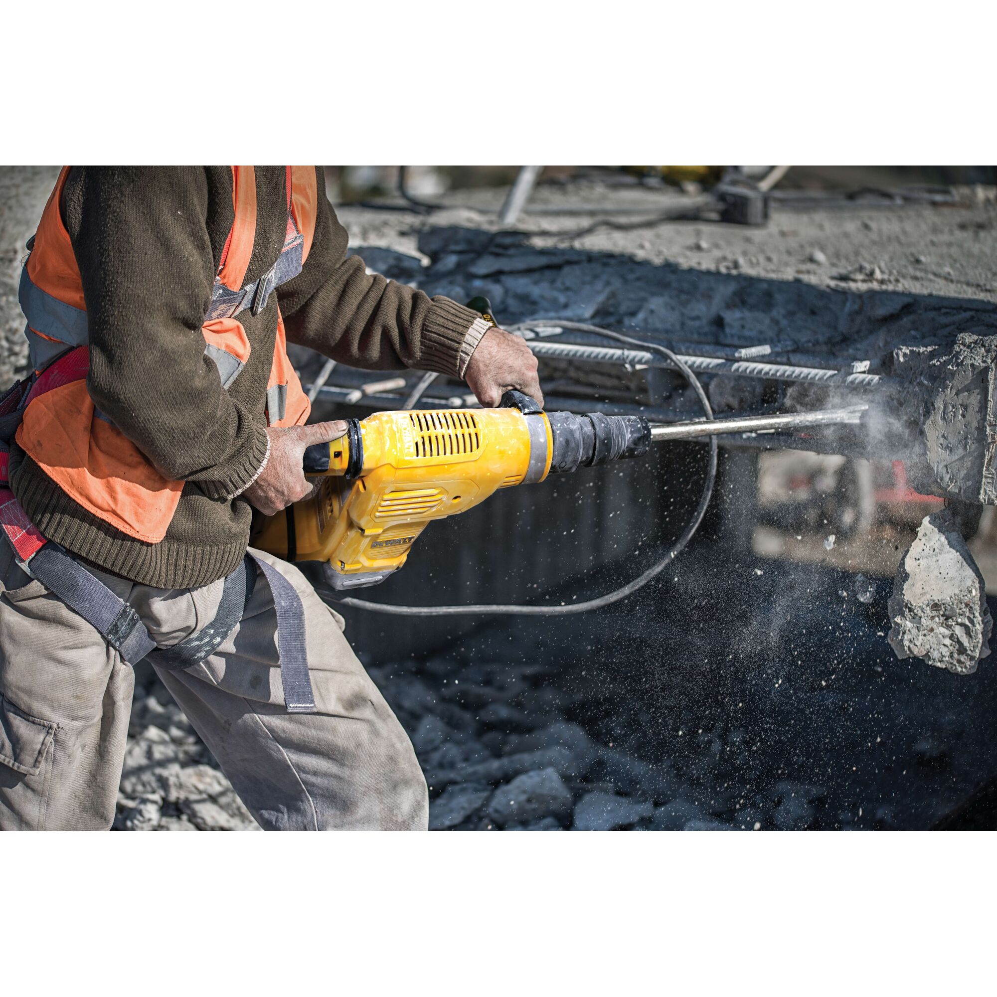 A person using a yellow DEWALT demolition hammer to break concrete at a construction site, wearing a safety harness and orange vest.