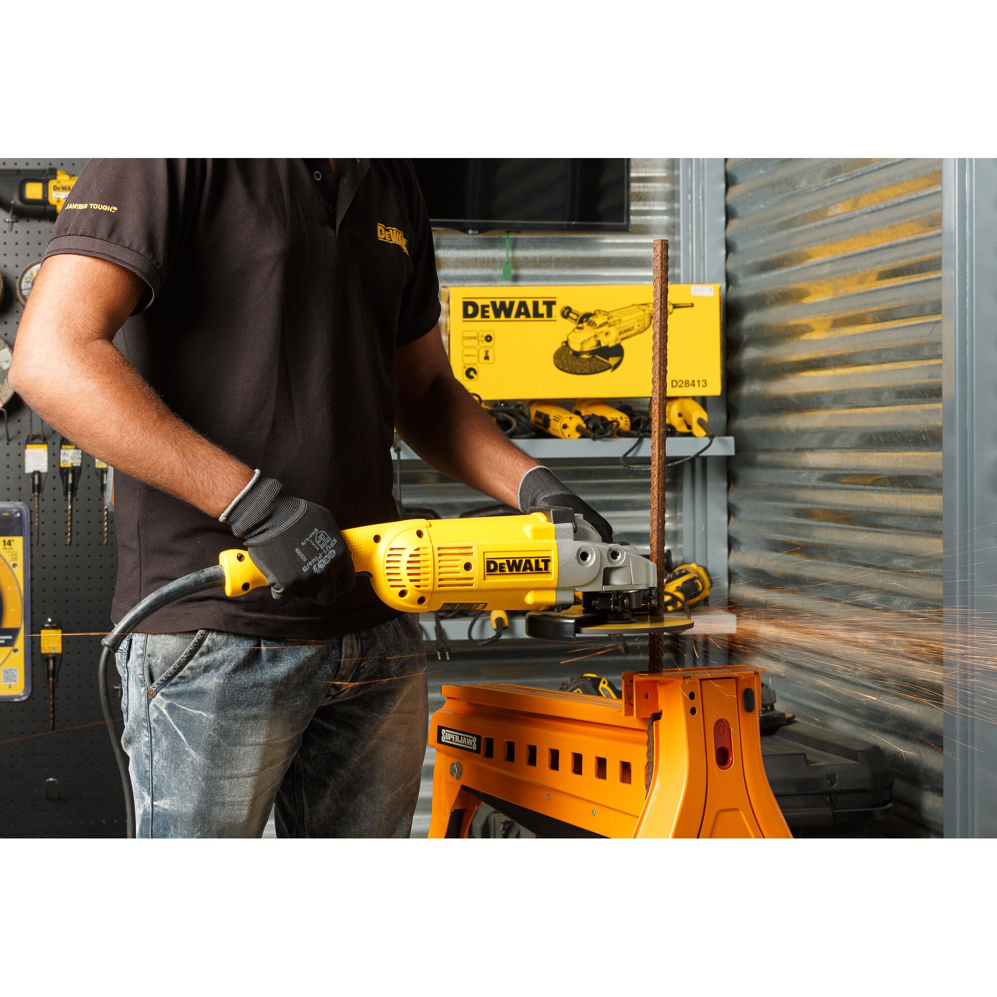 A person wearing gloves and a black shirt uses a yellow DEWALT D28413 angle grinder to cut a metal rod, producing sparks. The tool is resting on an orange workbench, with other DEWALT tools and a product box visible in the background.