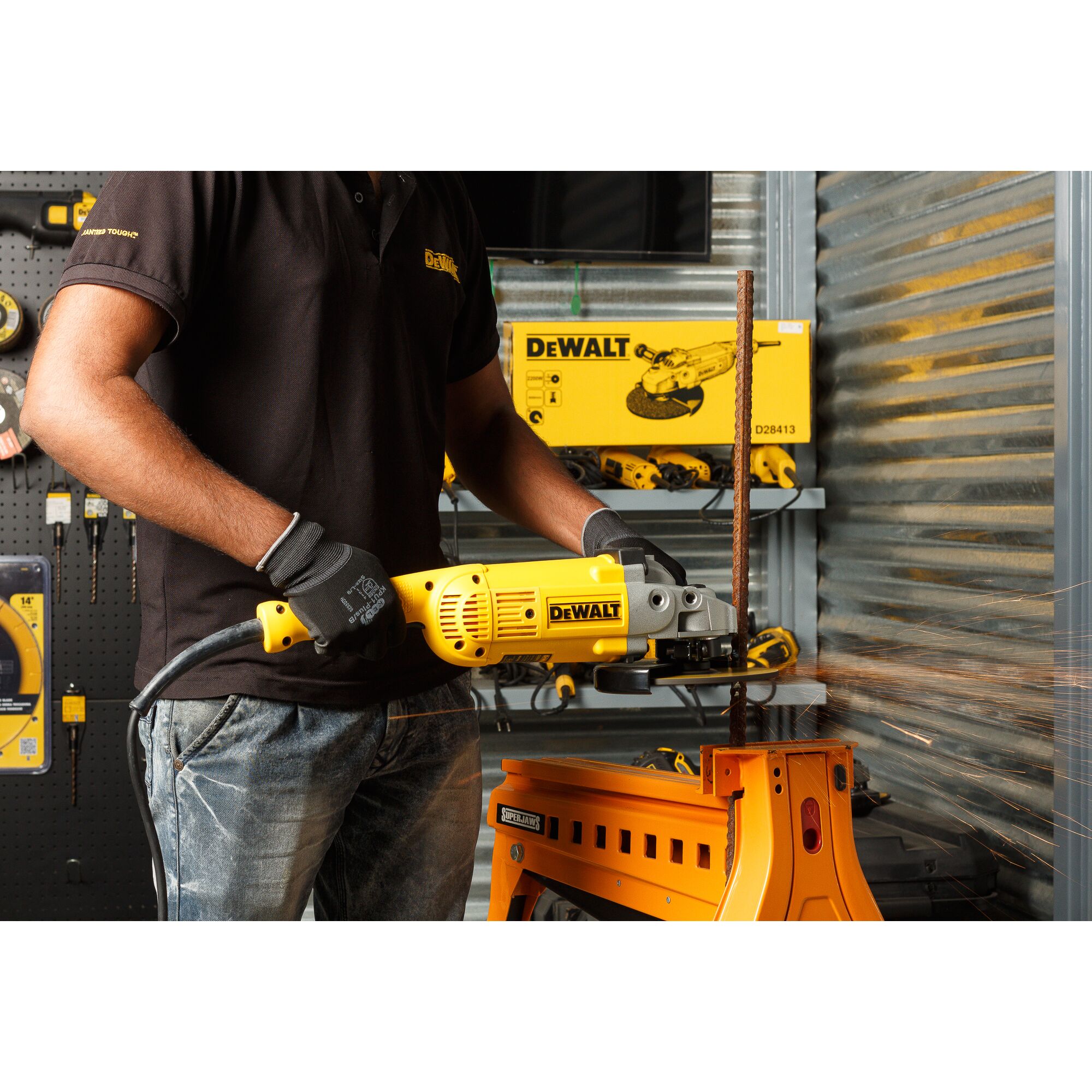 A person wearing gloves and a black shirt uses a yellow DEWALT angle grinder to cut a metal rod, producing sparks. The workspace includes a sturdy orange metal table and DEWALT products and tools displayed in the background.