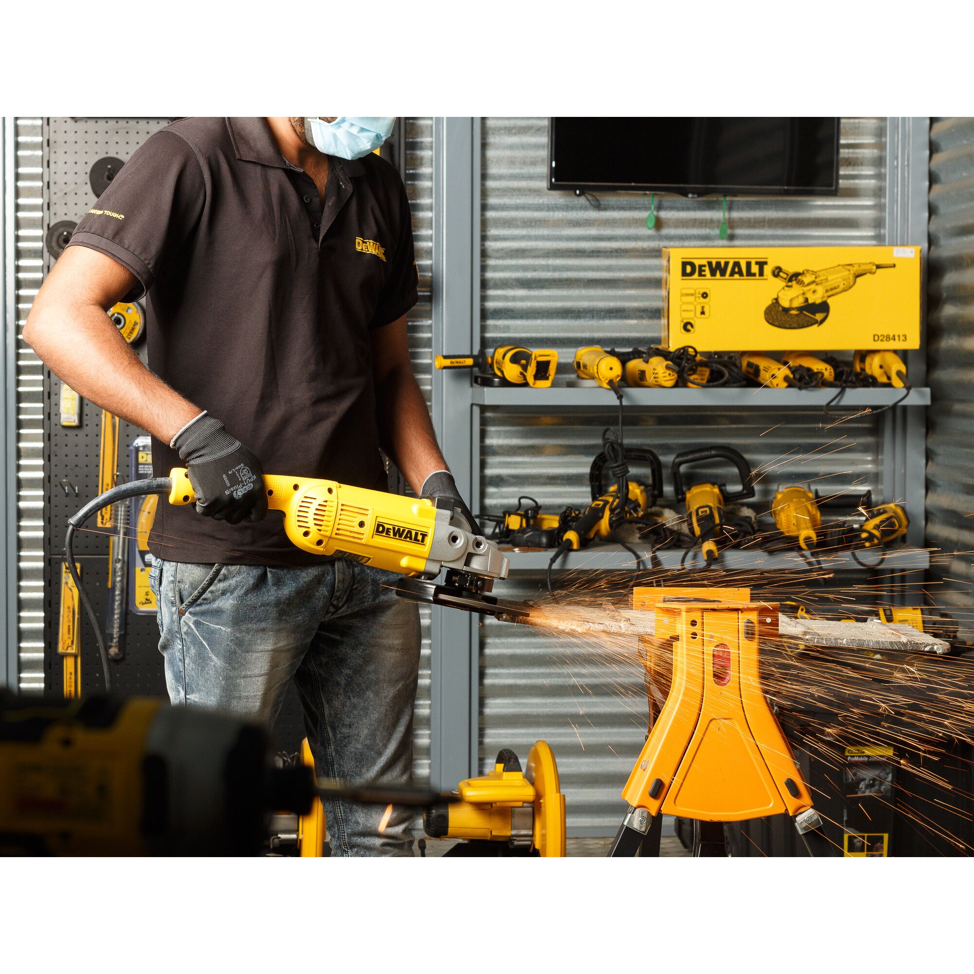 A person wearing a black shirt, gloves, and a face mask is using a DEWALT D28413 angle grinder to cut metal, creating sparks. The scene is set in a workshop with various DEWALT tools and equipment visible in the background.