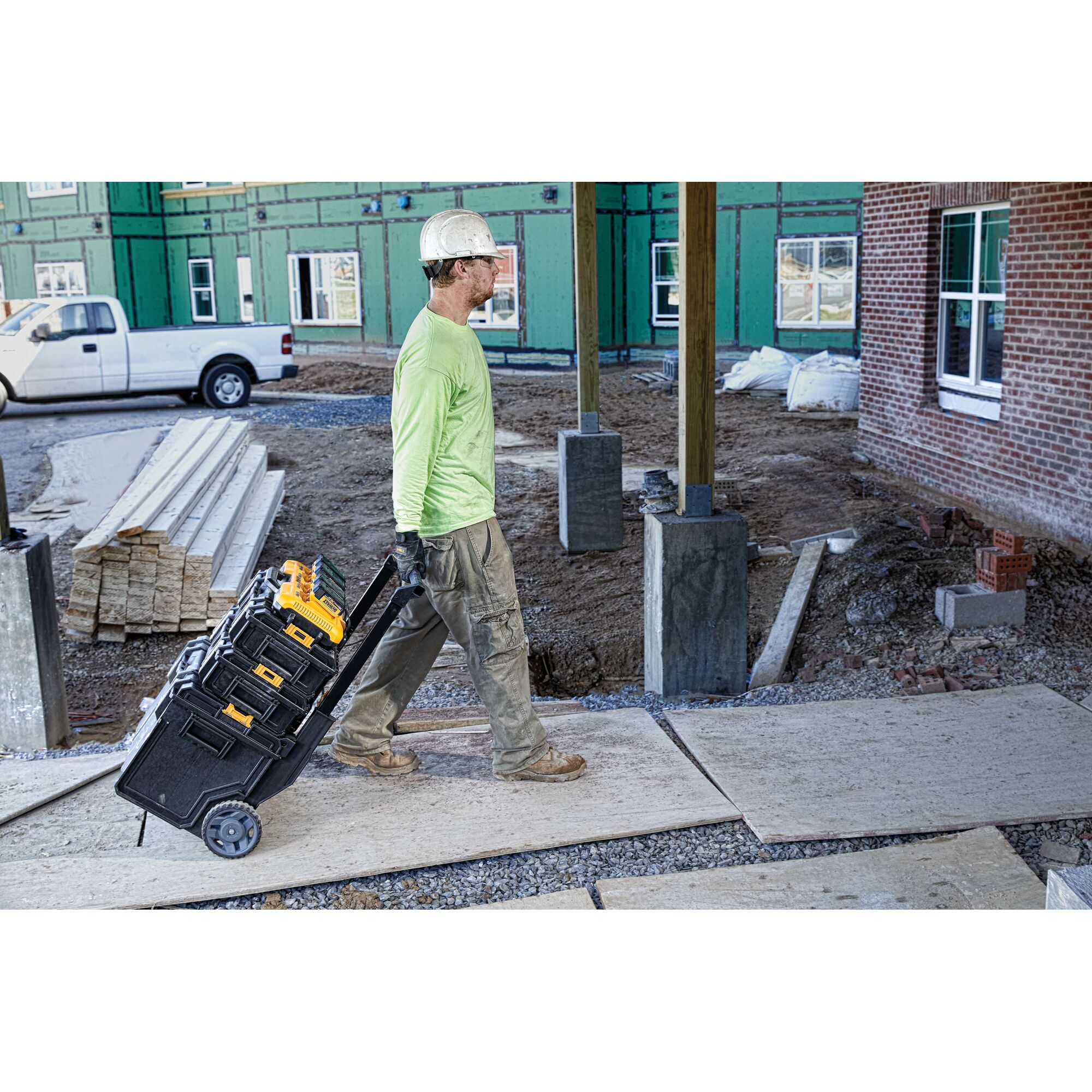 A construction worker wearing a hard hat and green shirt pulls a rolling cart stacked with black and yellow DEWALT toolboxes across a construction site.