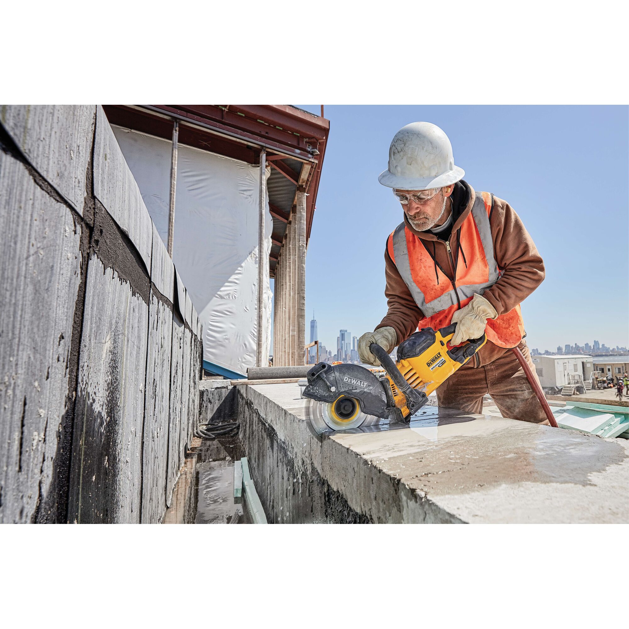 A worker wearing a safety vest, gloves, and helmet operates a yellow DEWALT concrete saw on a construction site. The background shows city buildings and construction materials.