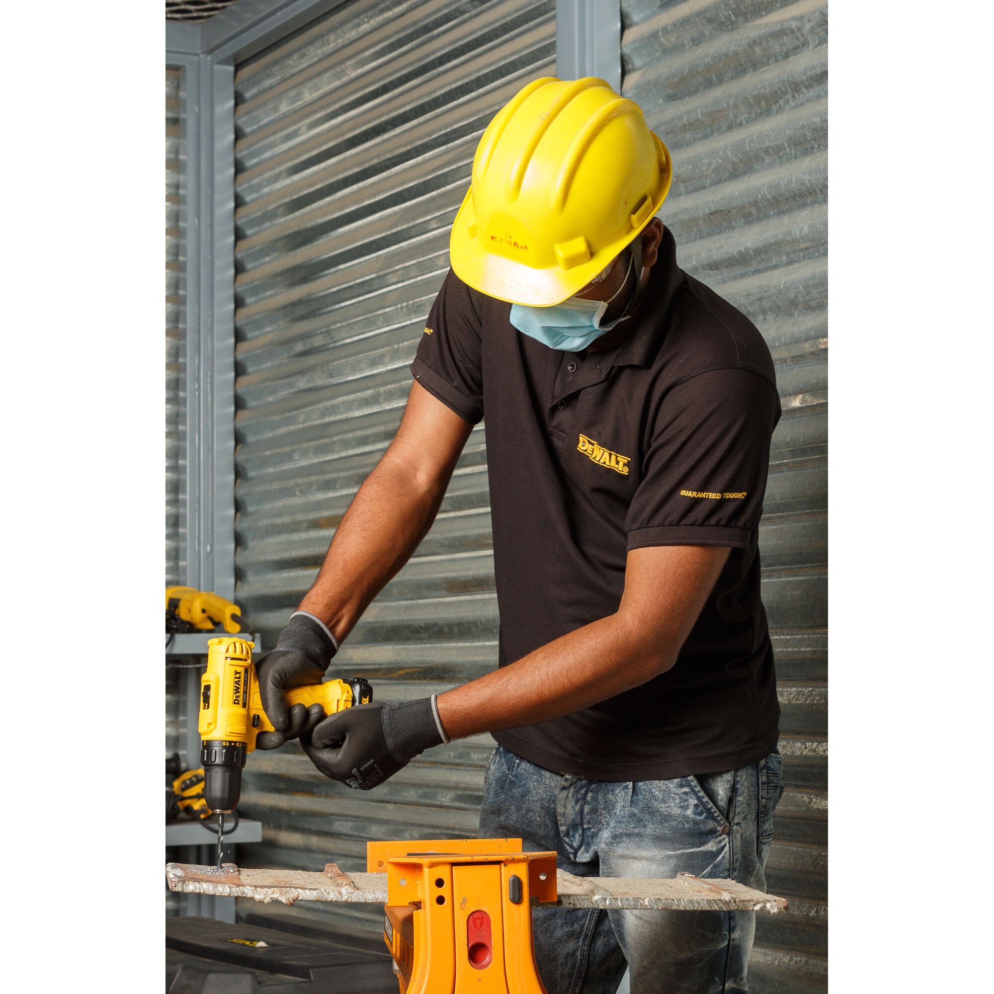 A person wearing a yellow hard hat, black DEWALT shirt, gloves, and a face mask is using a yellow DEWALT cordless drill (SKU: DCD700C2A) to drill into a piece of metal supported by an orange workbench in an industrial setting.