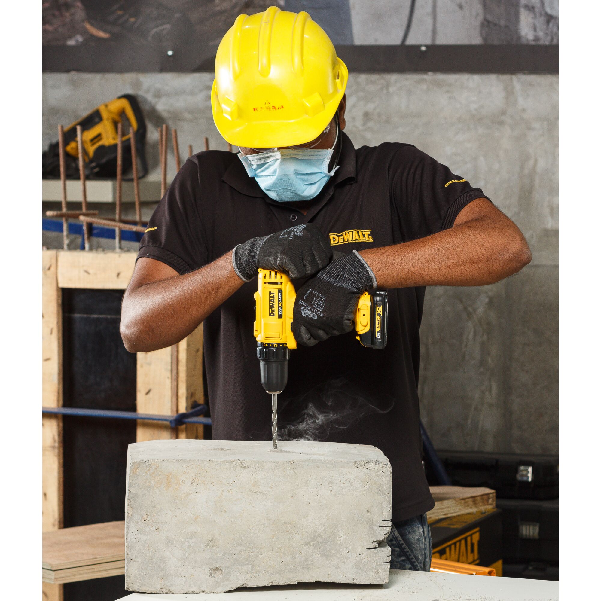 A person wearing a yellow hard hat and gloves uses a DEWALT cordless drill to drill into a concrete block. The scene takes place in an industrial or workshop setting.