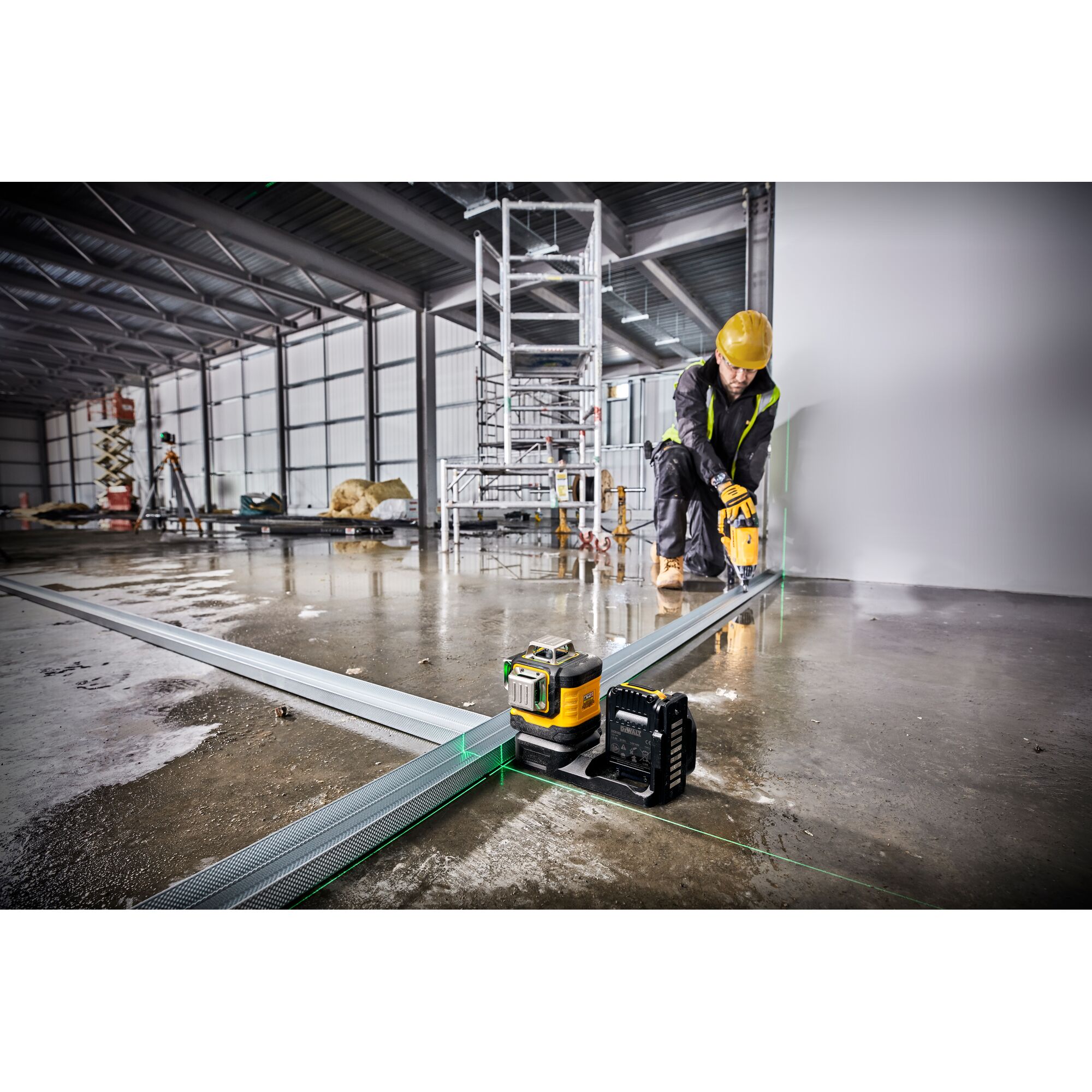 A DEWALT laser level tool (DCE089NG18) is placed on a construction site floor, projecting green laser lines along metal framing. A person wearing safety gear, including a helmet and vest, is working along the wall. The background shows scaffolding and industrial equipment.