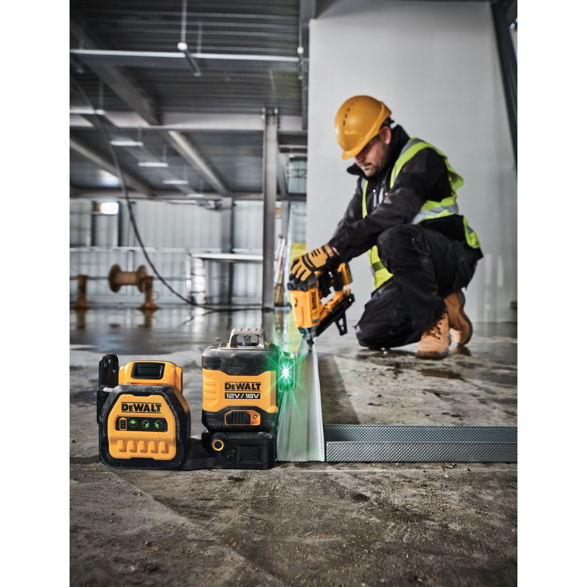 A DEWALT 12V/18V green cross line laser level is shown on a construction site floor alongside another DEWALT measuring tool. In the background, a construction worker wearing personal protective equipment including a hard hat, gloves, and high-visibility vest, is kneeling and using a DEWALT power tool on a metal frame.