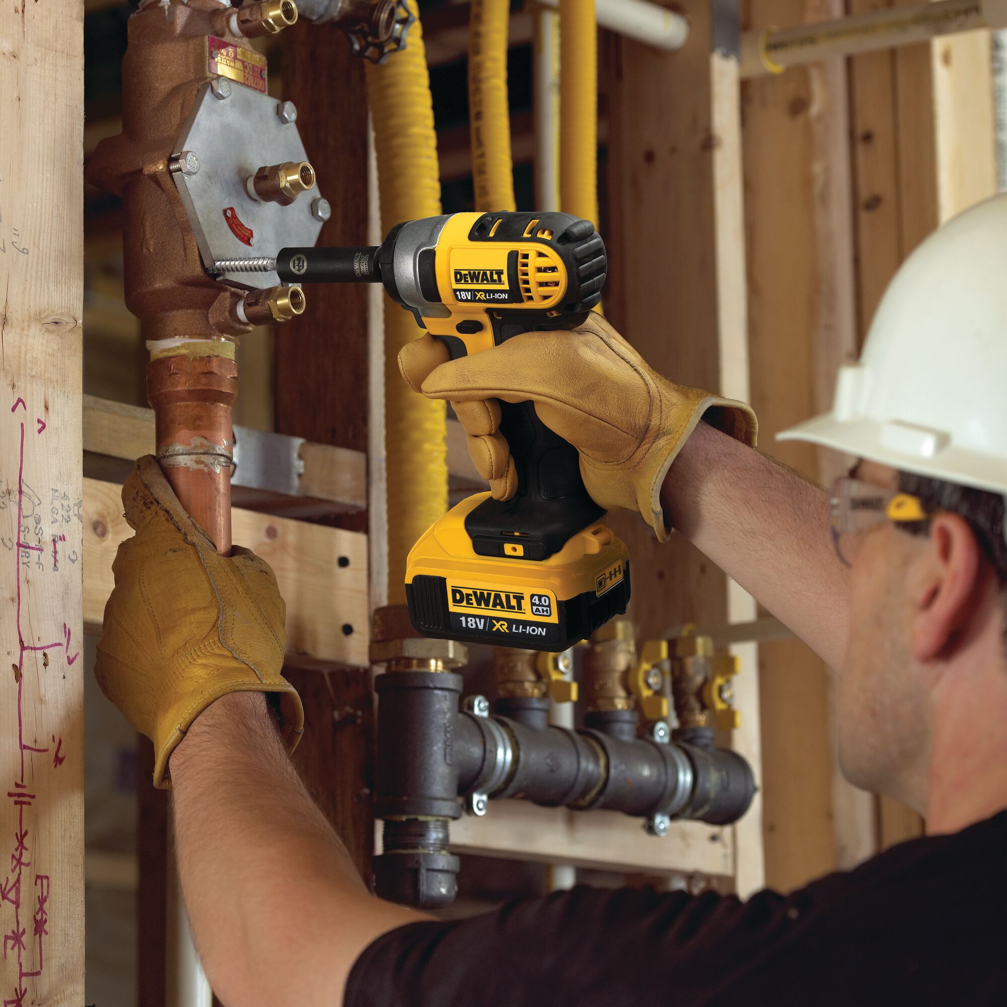A person wearing work gloves and a hard hat uses a DEWALT DCF880N 18V XL Li-Ion cordless impact wrench on a metal pipe in a construction setting.