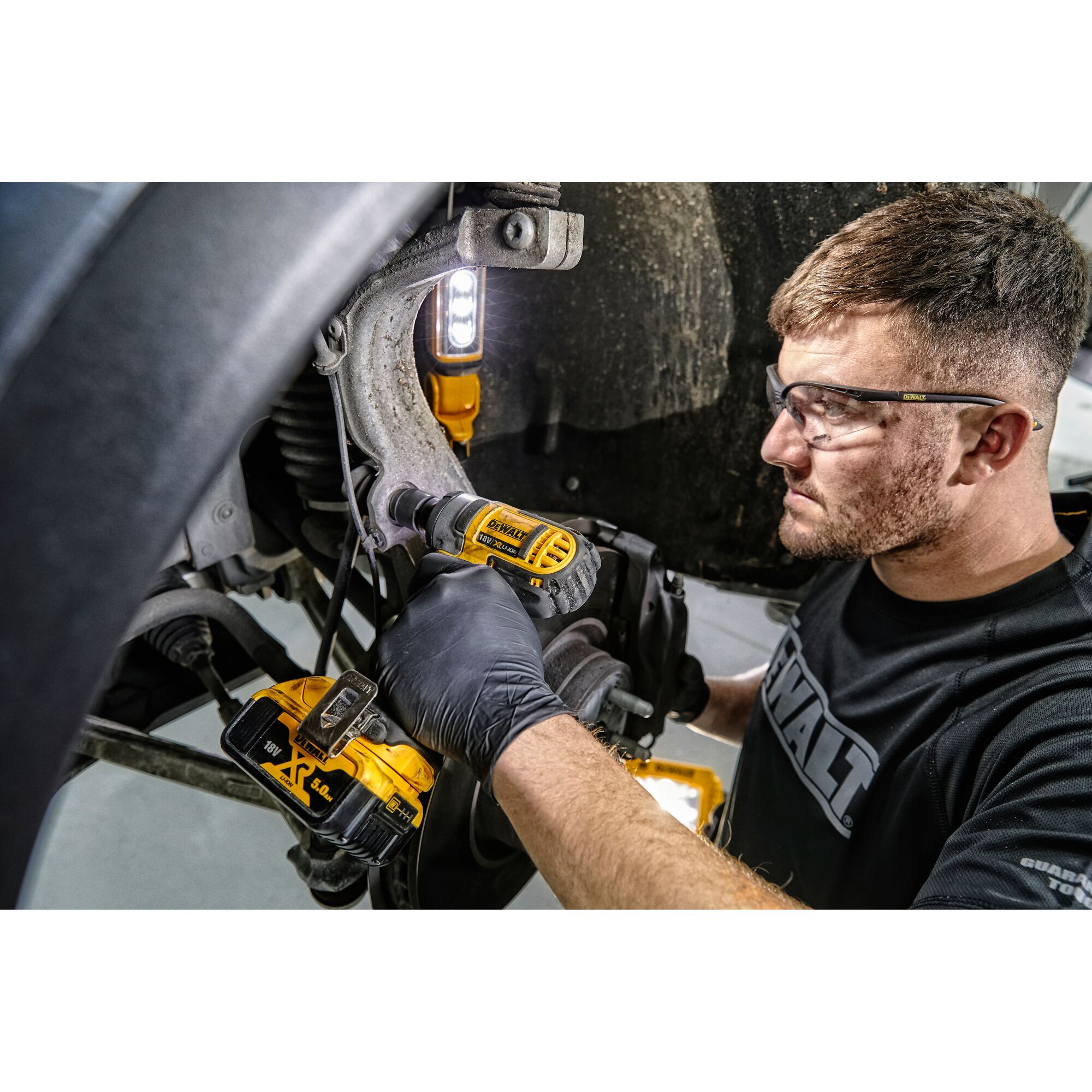 A person wearing gloves is using a DEWALT DCF880N cordless impact wrench to perform maintenance on a vehicle suspension component. The DEWALT tool is yellow and black, and a portable LED work light illuminates the workspace. The person's face is not visible.