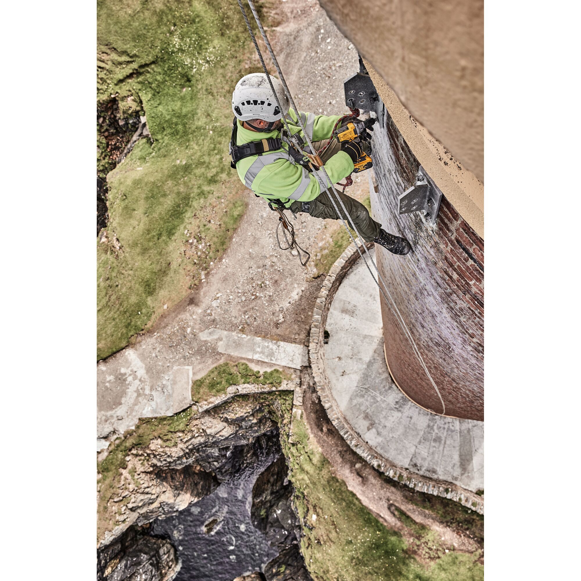 A worker wearing safety gear, including a helmet and harness, is using a DEWALT power tool while suspended by ropes against the side of a brick tower in an outdoor setting.