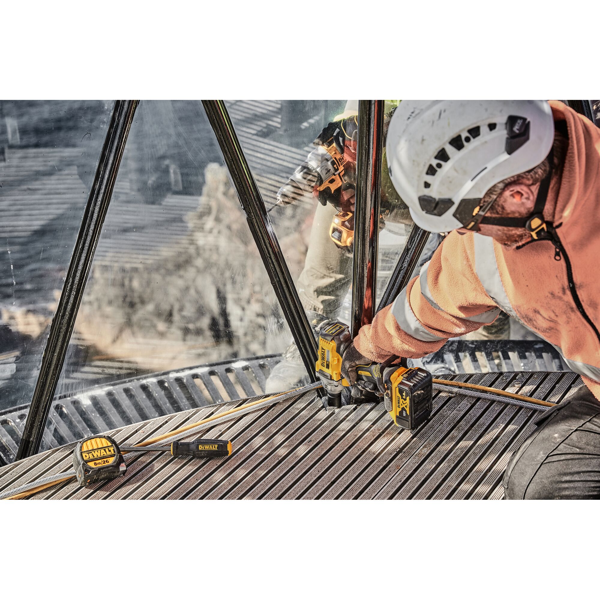 A person wearing a white safety helmet and orange workwear is using a DEWALT cordless impact wrench on a metal structure. The DEWALT DCF894P2 tool and battery are visible, along with a DEWALT measuring tape and screwdriver lying nearby.