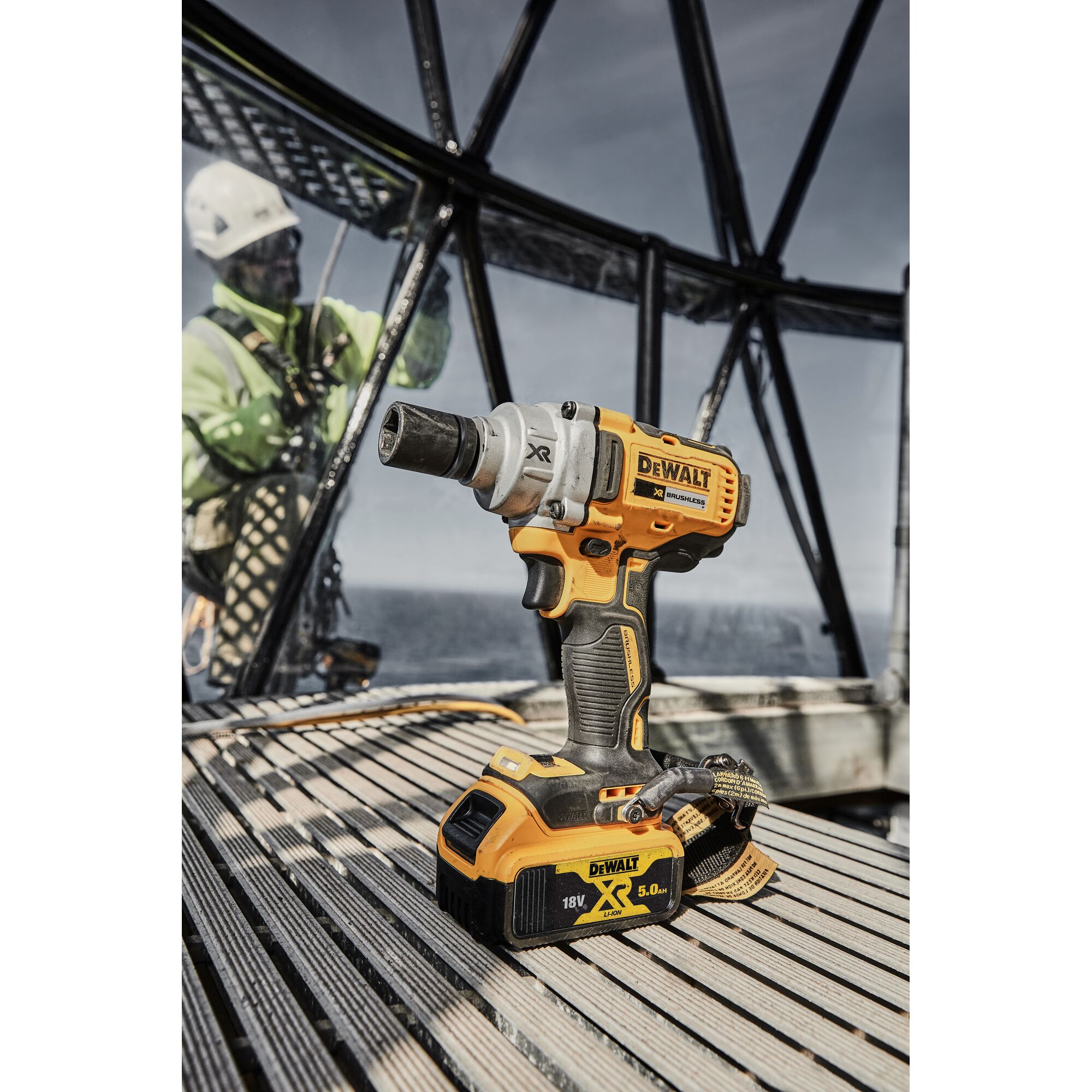 A DEWALT DCF894P2 cordless impact wrench with an 18V XR Lithium-ion 5.0Ah battery is placed on a wooden surface. In the background, there is a person in safety gear working inside a metal structure with a view of the sea.