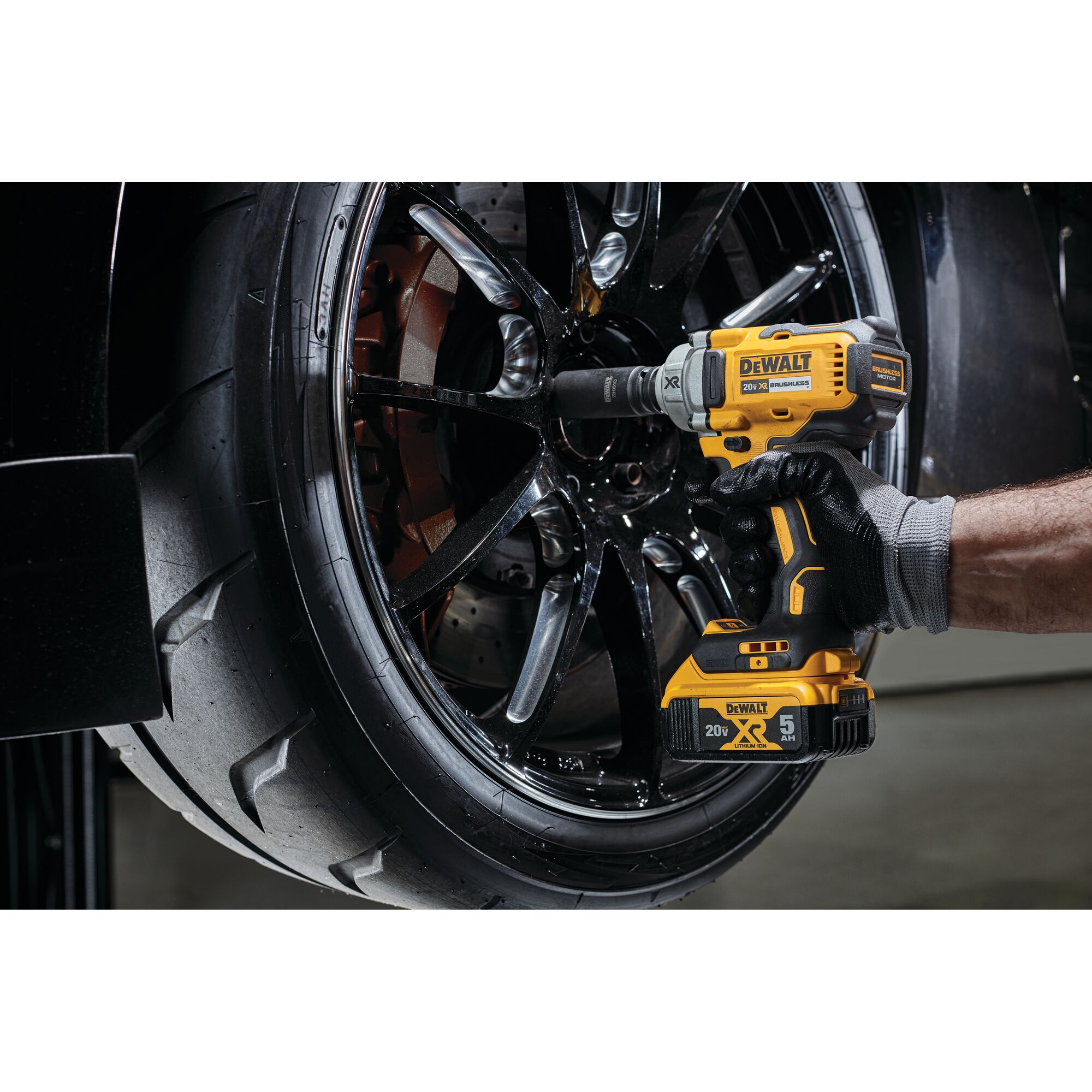 A gloved hand uses a yellow and black DEWALT cordless impact wrench to tighten or loosen bolts on a black car wheel.