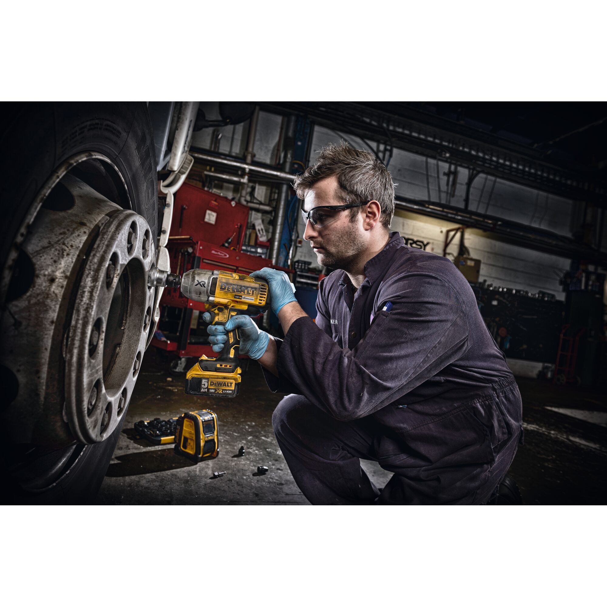 Person wearing a dark jumpsuit and blue gloves using a yellow DEWALT power tool to work on the wheel of a large vehicle. The scene is set in a workshop with various equipment visible in the background.