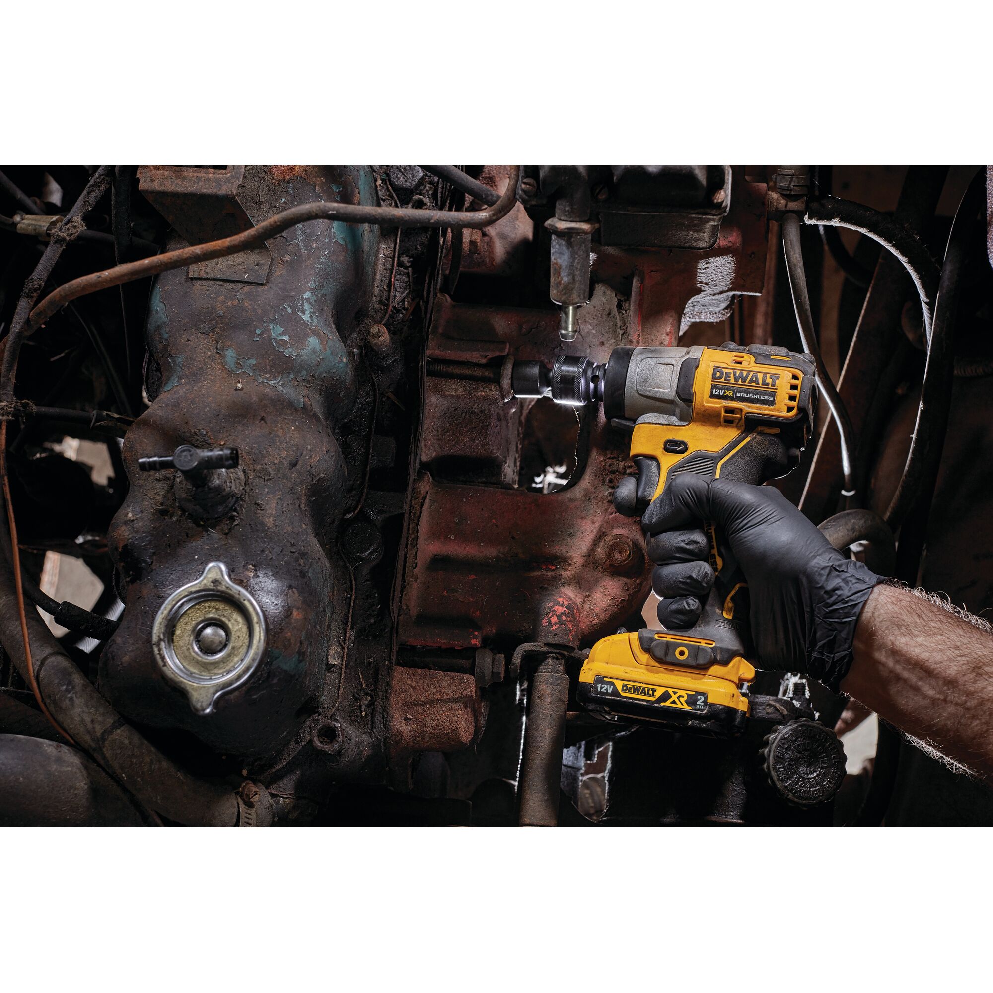 A person wearing a black glove uses a DEWALT DCF902D2 cordless impact wrench to work on a part of an engine or machinery. The tool is yellow and black and is being applied to a rusty metal component.