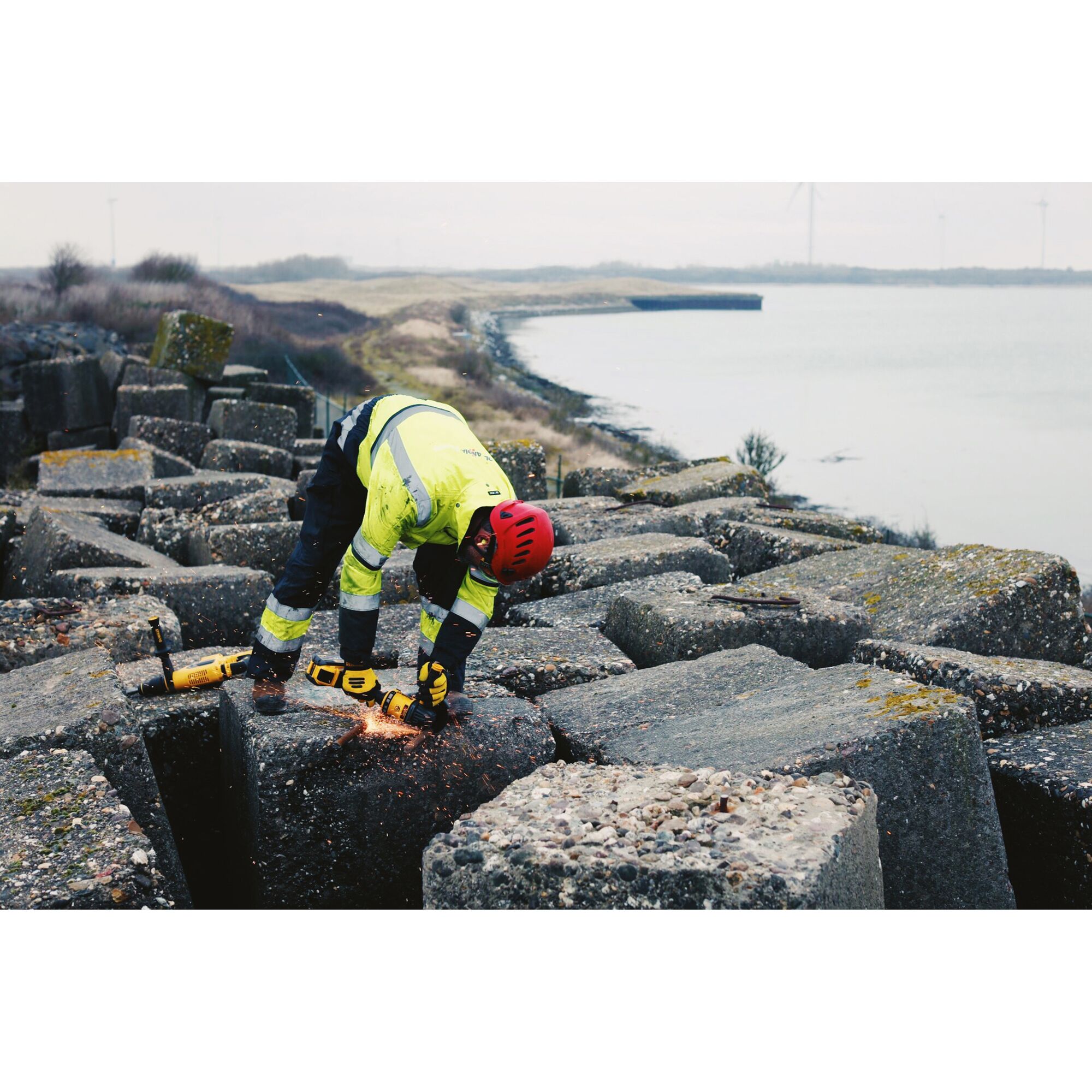 A person wearing a safety helmet and high-visibility jacket uses a yellow DEWALT DCG414N angle grinder to cut into a large concrete block outdoors near a body of water, with sparks visible during operation.