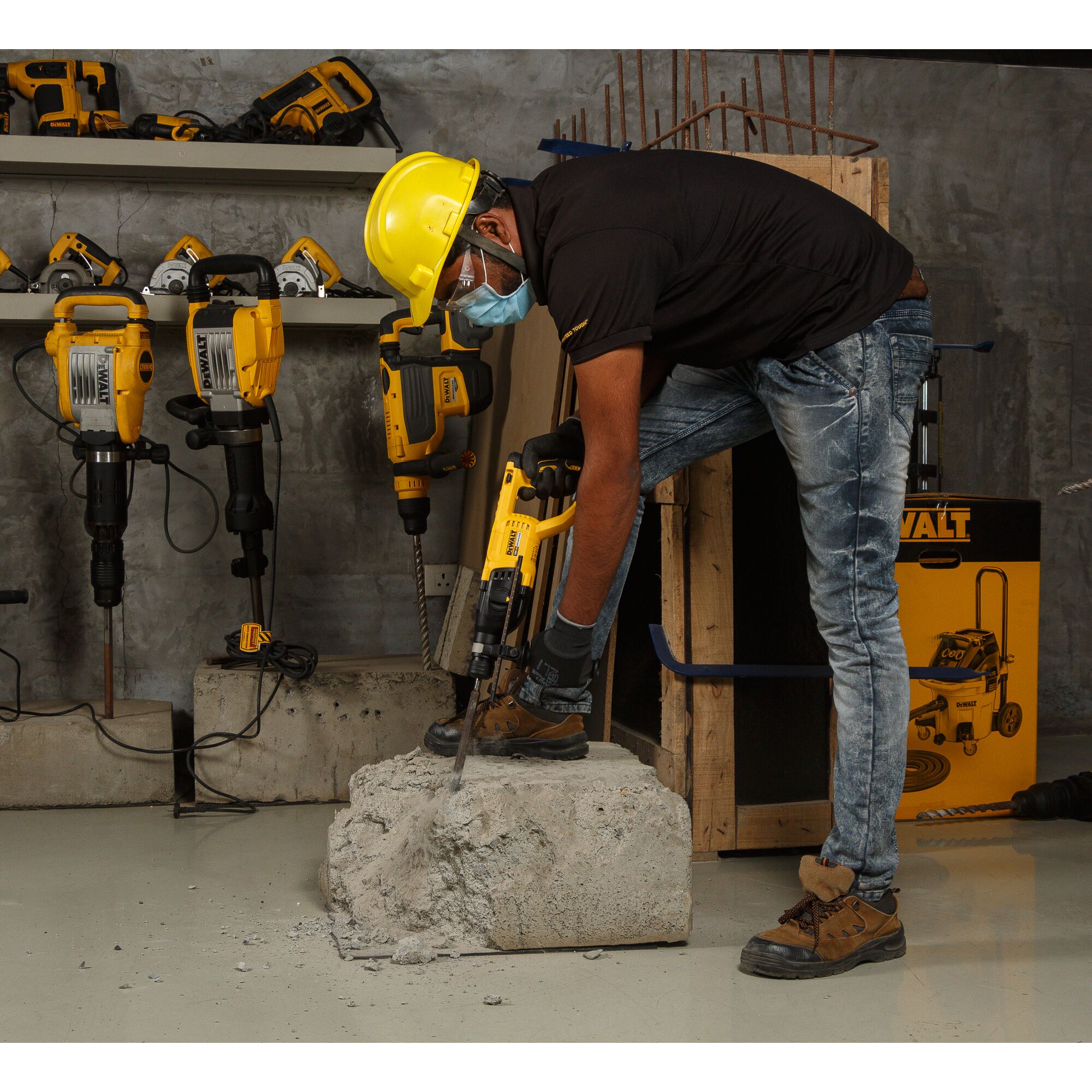 A worker wearing a yellow safety helmet, face mask, gloves, and work boots is using a DEWALT rotary hammer drill to break a concrete block in an industrial setting. Several DEWALT power tools are displayed on shelves in the background.