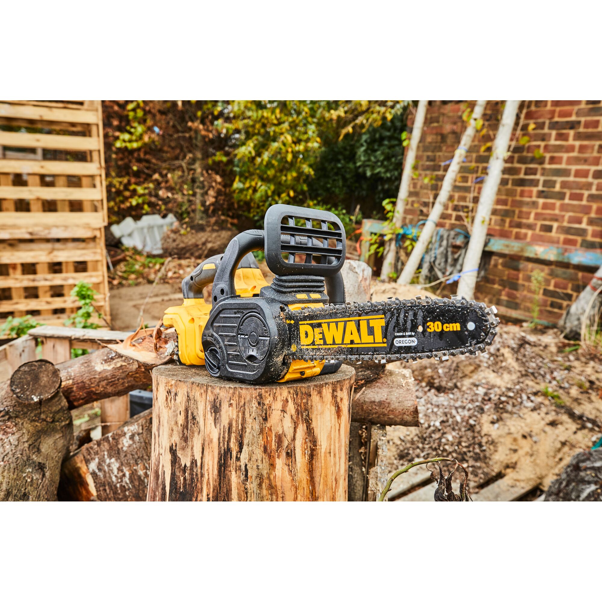 A DEWALT DCM565N 30 cm cordless chainsaw resting on a tree stump in an outdoor setting, surrounded by logs and a wooden pallet.