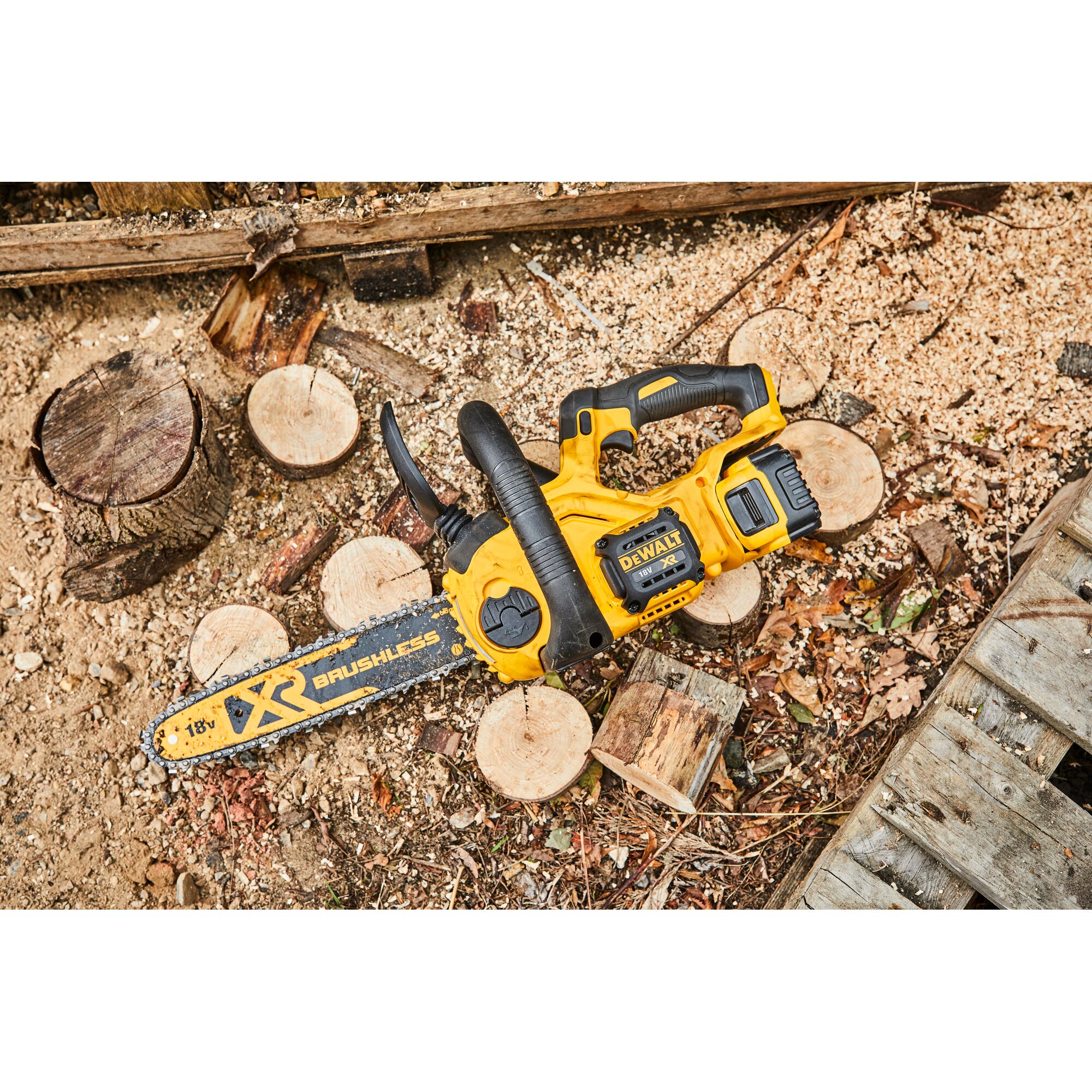 A DEWALT DCM565N 18V XR brushless cordless chainsaw placed on the ground next to cut wooden logs and sawdust in an outdoor setting.