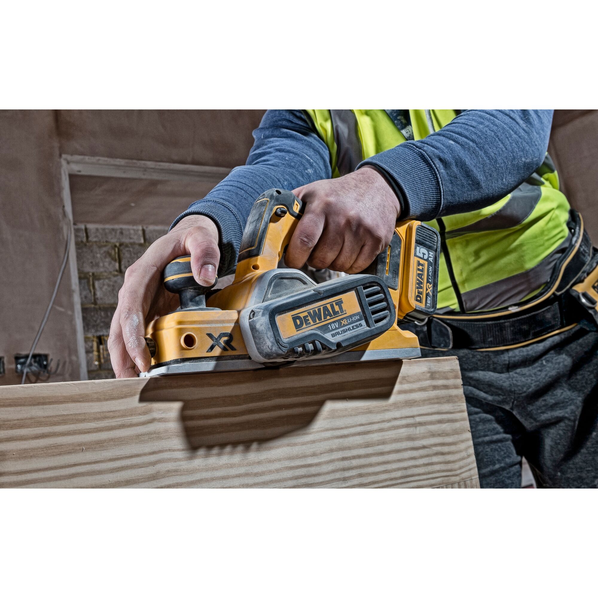 Worker uses a DEWALT cordless planer on a wooden plank in a construction setting.