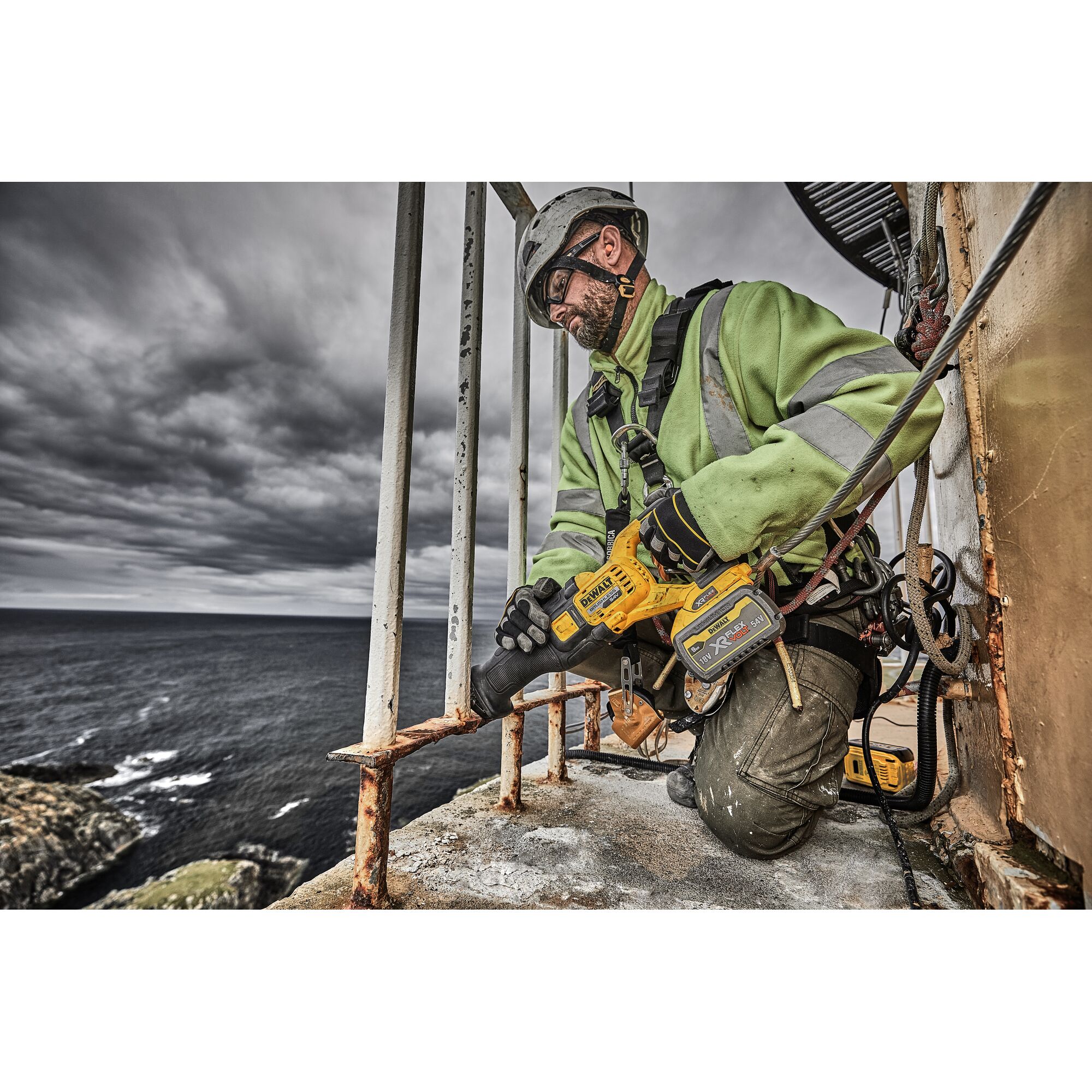 A worker wearing a safety helmet and high-visibility jacket uses a DEWALT DCS389X2 cordless reciprocating saw to cut through a rusty metal railing outdoors near the coast, with dark clouds overhead.
