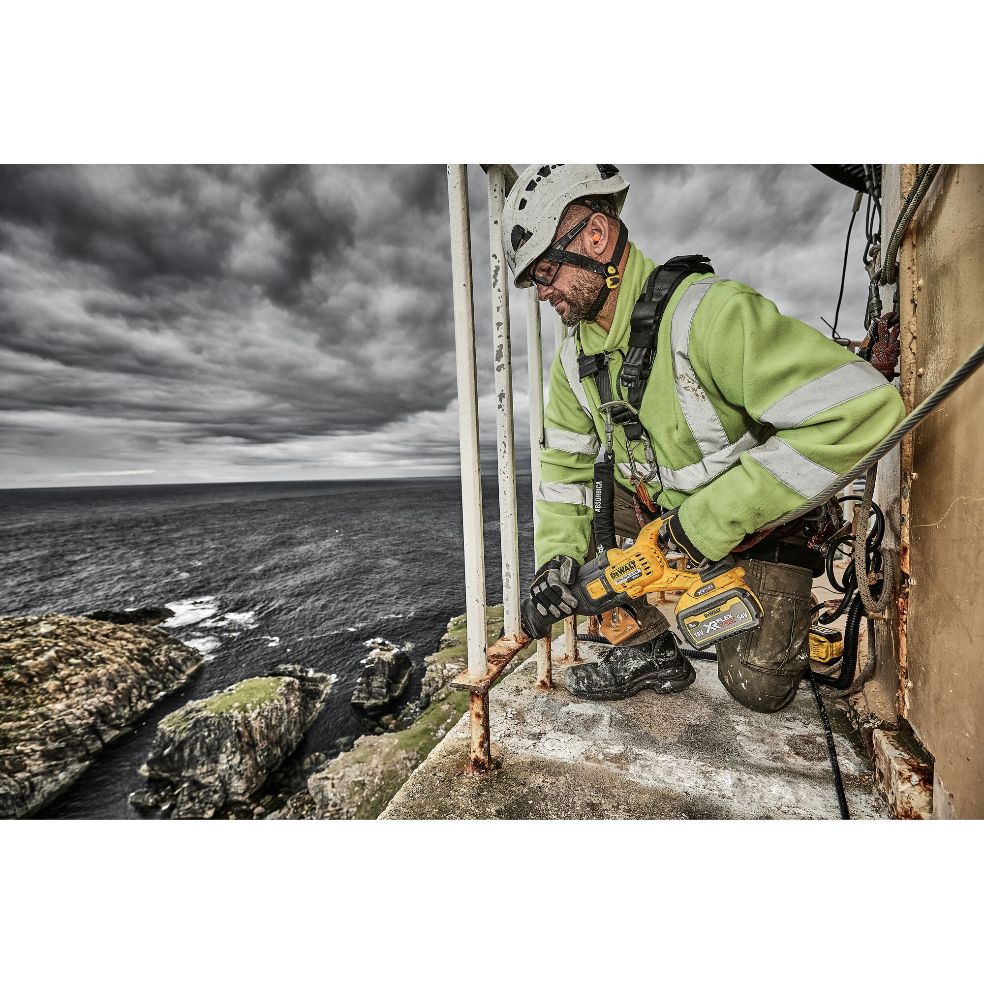 A person wearing safety gear and a green reflective jacket is using a DEWALT cordless power tool to cut metal rods on an outdoor platform near the edge of a cliff, with an ocean and rocky coastline in the background.