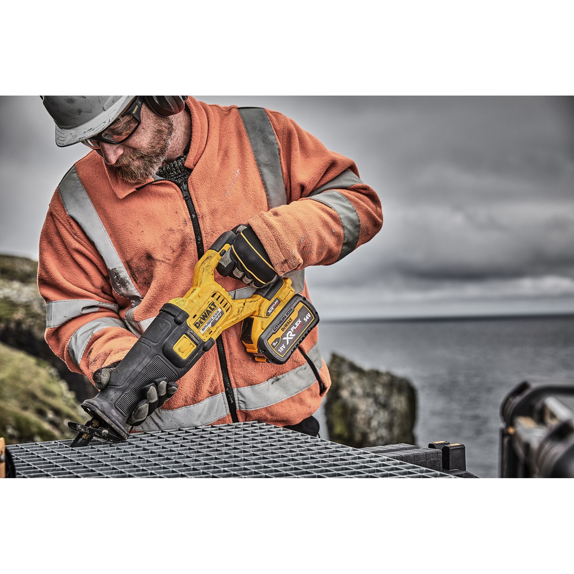 A person wearing an orange high-visibility jacket and gloves is holding a DEWALT DCS389X2 cordless reciprocating saw while working outdoors on a metal surface, with a blurred background of landscape and water.