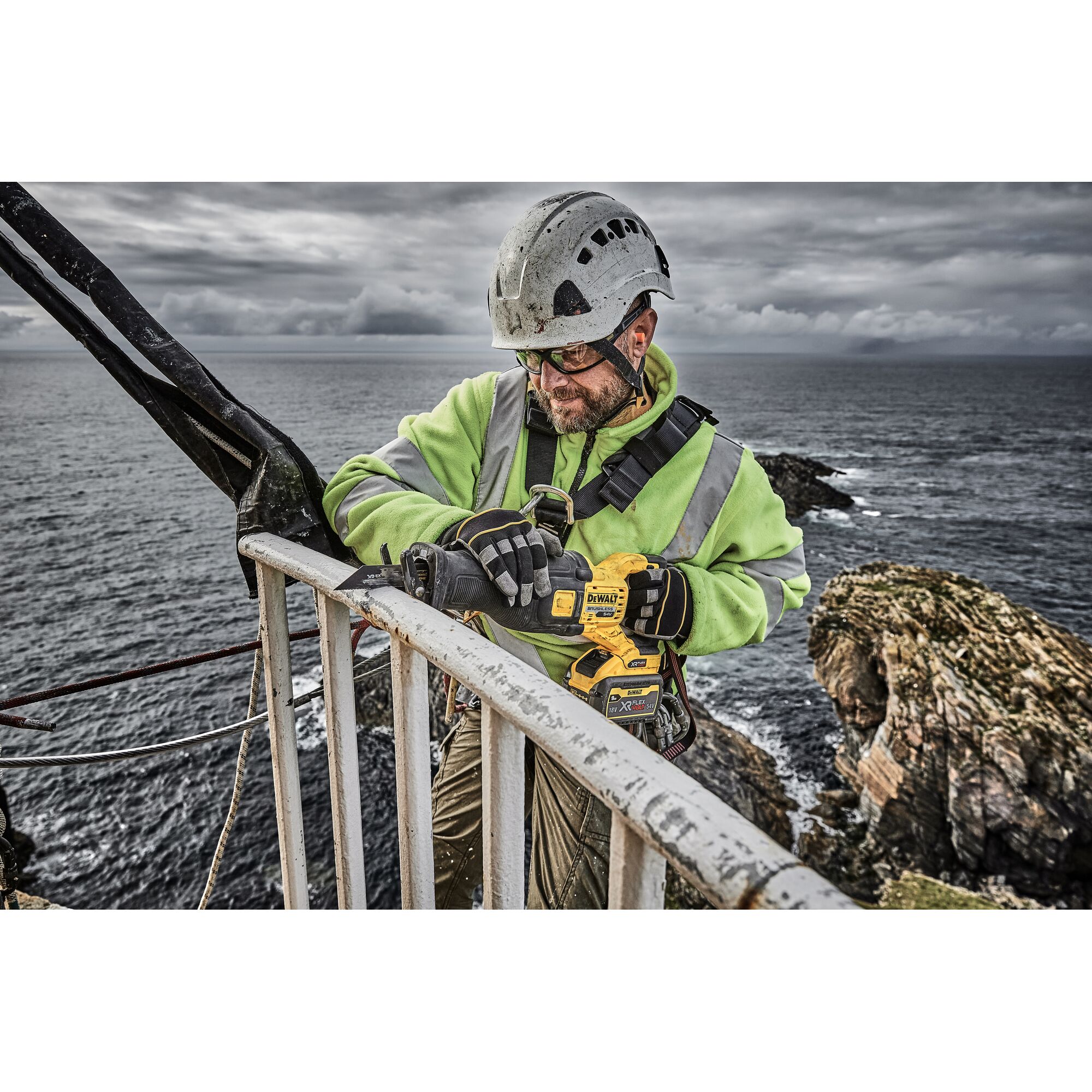 A worker wearing a safety helmet, gloves, and a high-visibility jacket is using a DEWALT DCS389X2 power tool on a metal railing outdoors, with rocky cliffs and the ocean in the background.