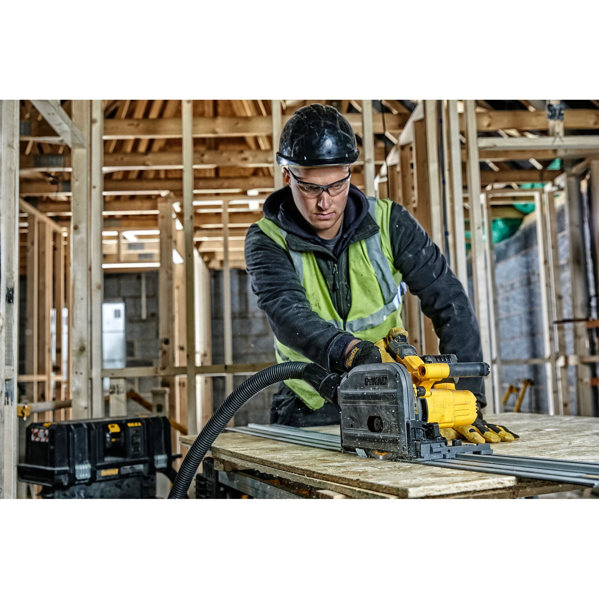 Worker uses DEWALT DCS520NT track saw to cut wood in a building under construction. Safety gear and tools are visible.