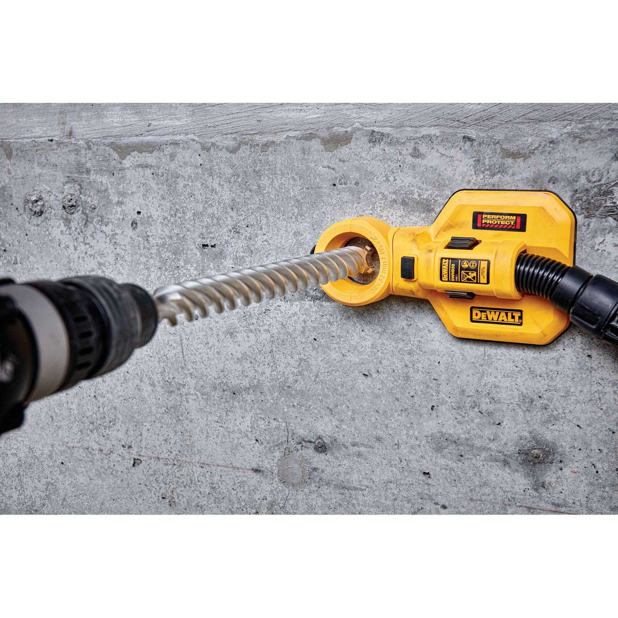 A DEWALT dust extraction attachment is being used with a rotary hammer drill to drill into concrete. The yellow DEWALT device is positioned against the wall to collect dust while drilling.