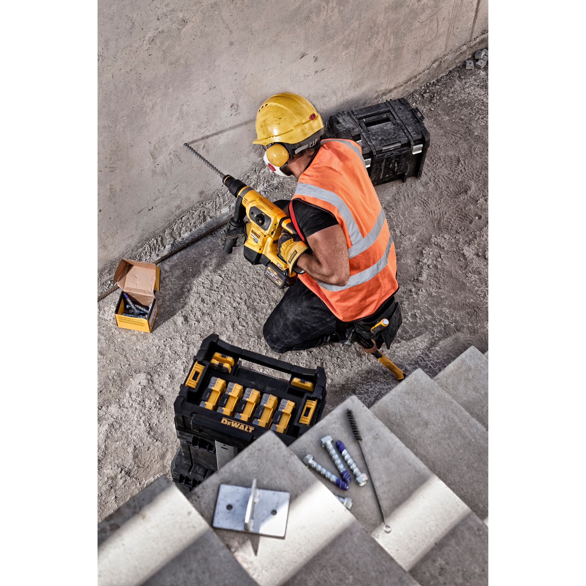 A construction worker wearing a yellow hard hat, orange safety vest, and gloves is using a DEWALT power drill to work on a concrete wall. There are DEWALT toolboxes and a box of drill bits nearby, as well as metal anchors and tools laid out on concrete steps.