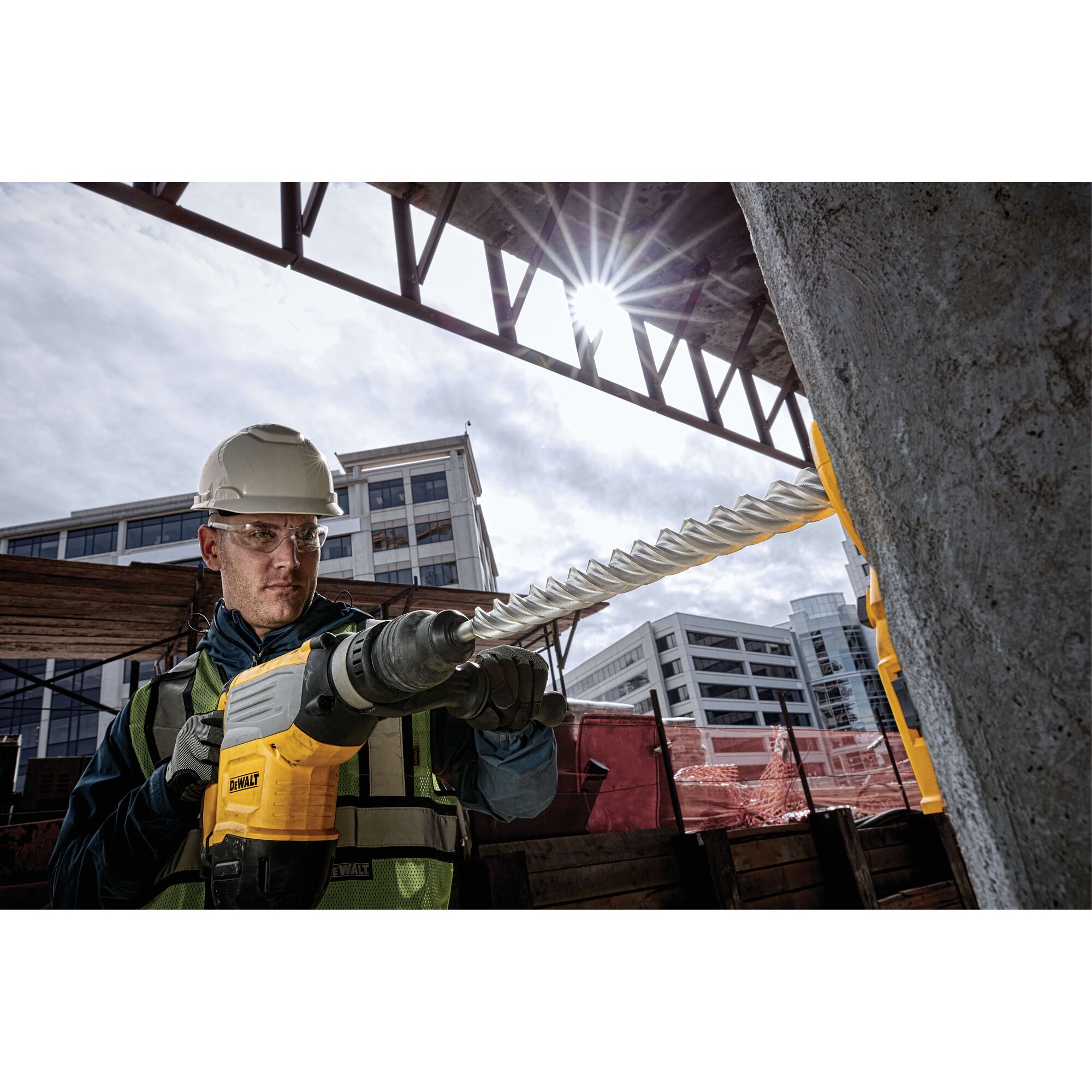 A construction worker using a DEWALT power drill with a long spiral bit to drill into a concrete pillar at an outdoor construction site. The worker is wearing a safety helmet and vest. Modern buildings are visible in the background.