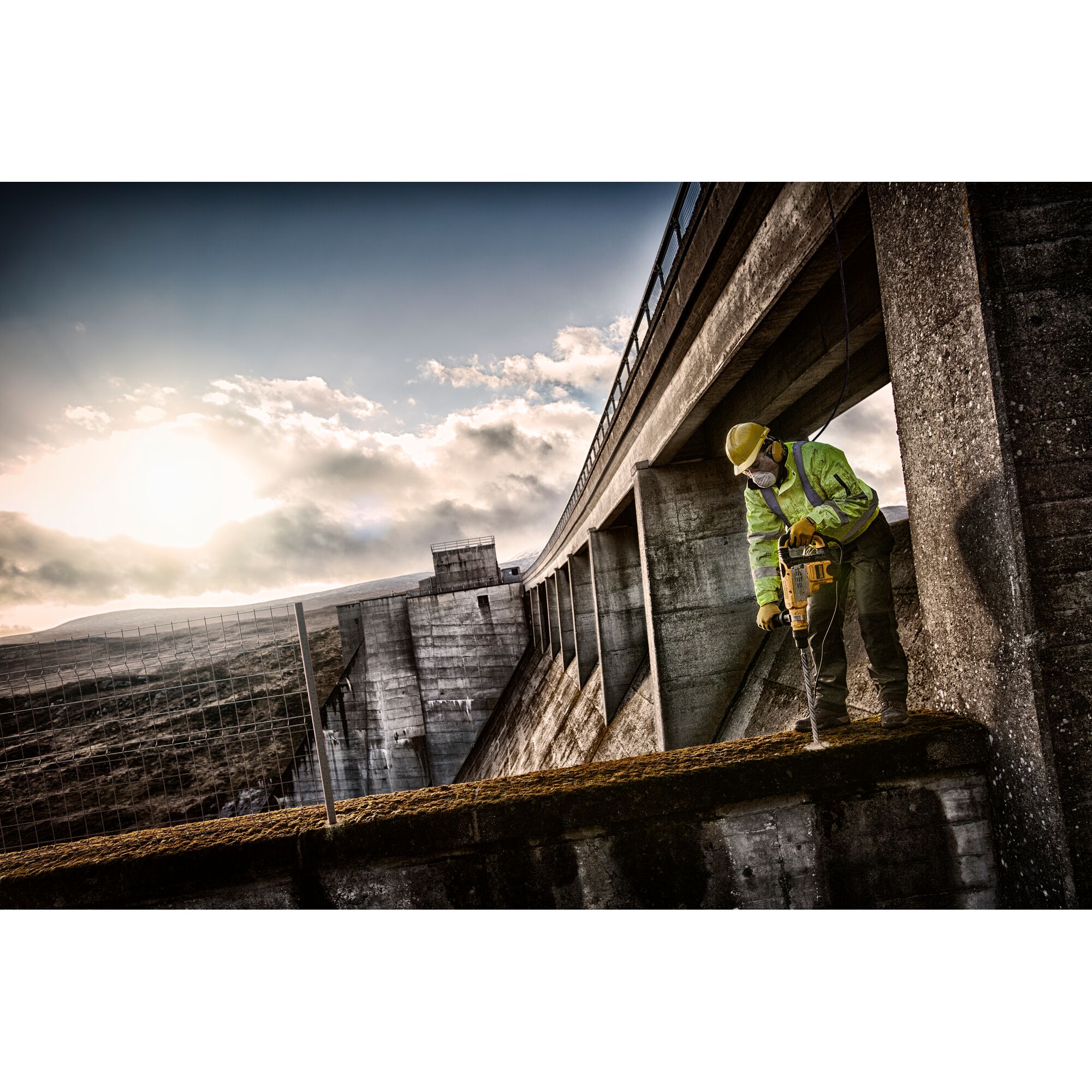 A worker wearing safety gear uses a DEWALT power tool to drill into concrete at an industrial construction site, with a large dam structure and dramatic sky in the background.