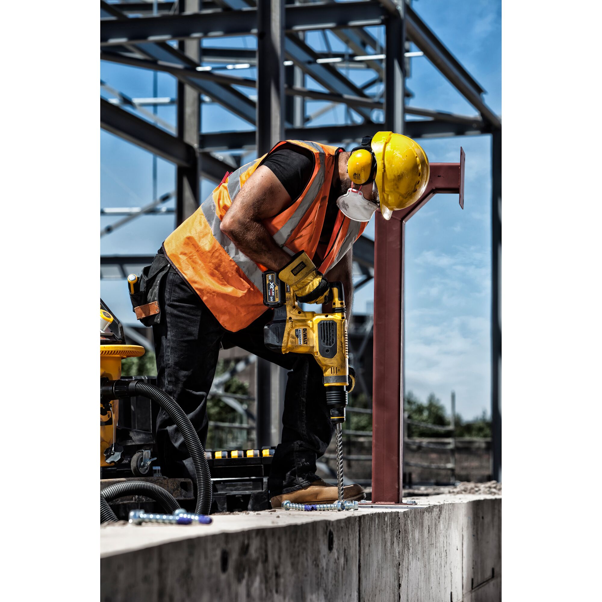 A construction worker wearing a yellow hard hat, orange safety vest, and face mask is using a DEWALT power drill to drill into a concrete surface at an outdoor construction site. Steel framework is visible in the background.