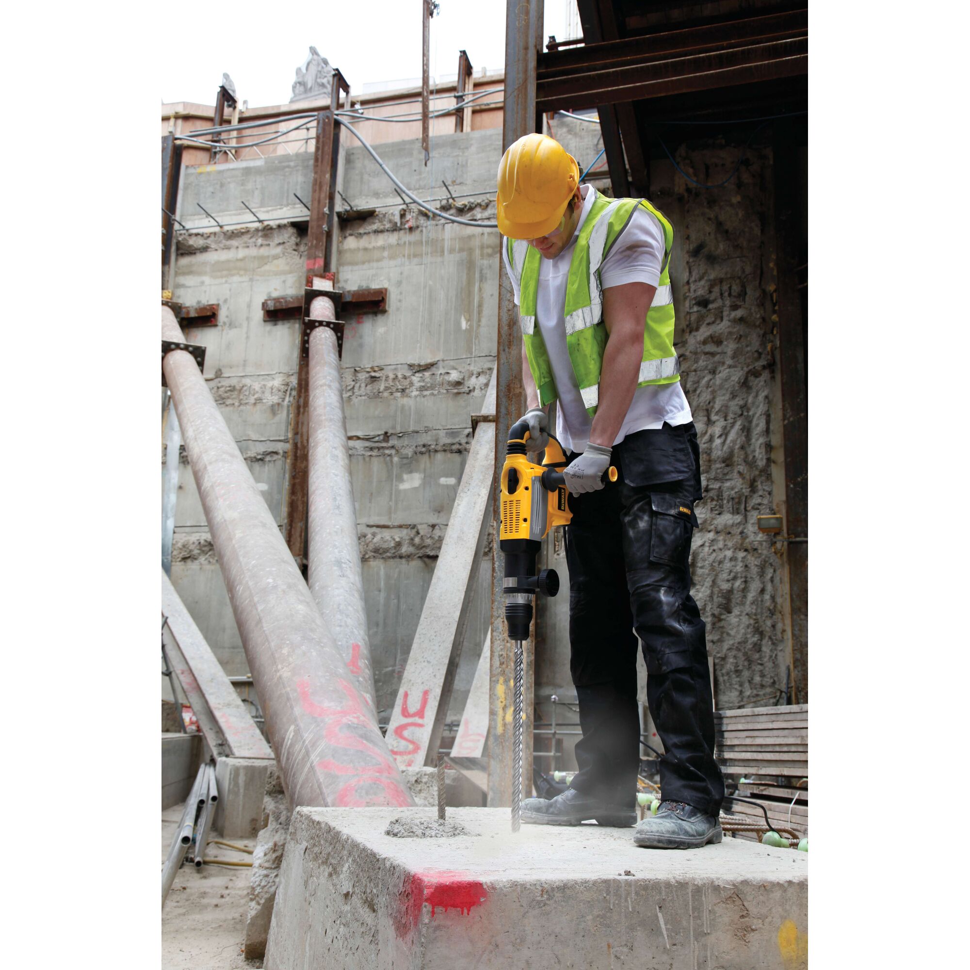 A construction worker wearing a yellow hard hat and reflective safety vest is using a DEWALT rotary hammer drill on a concrete block at a construction site.