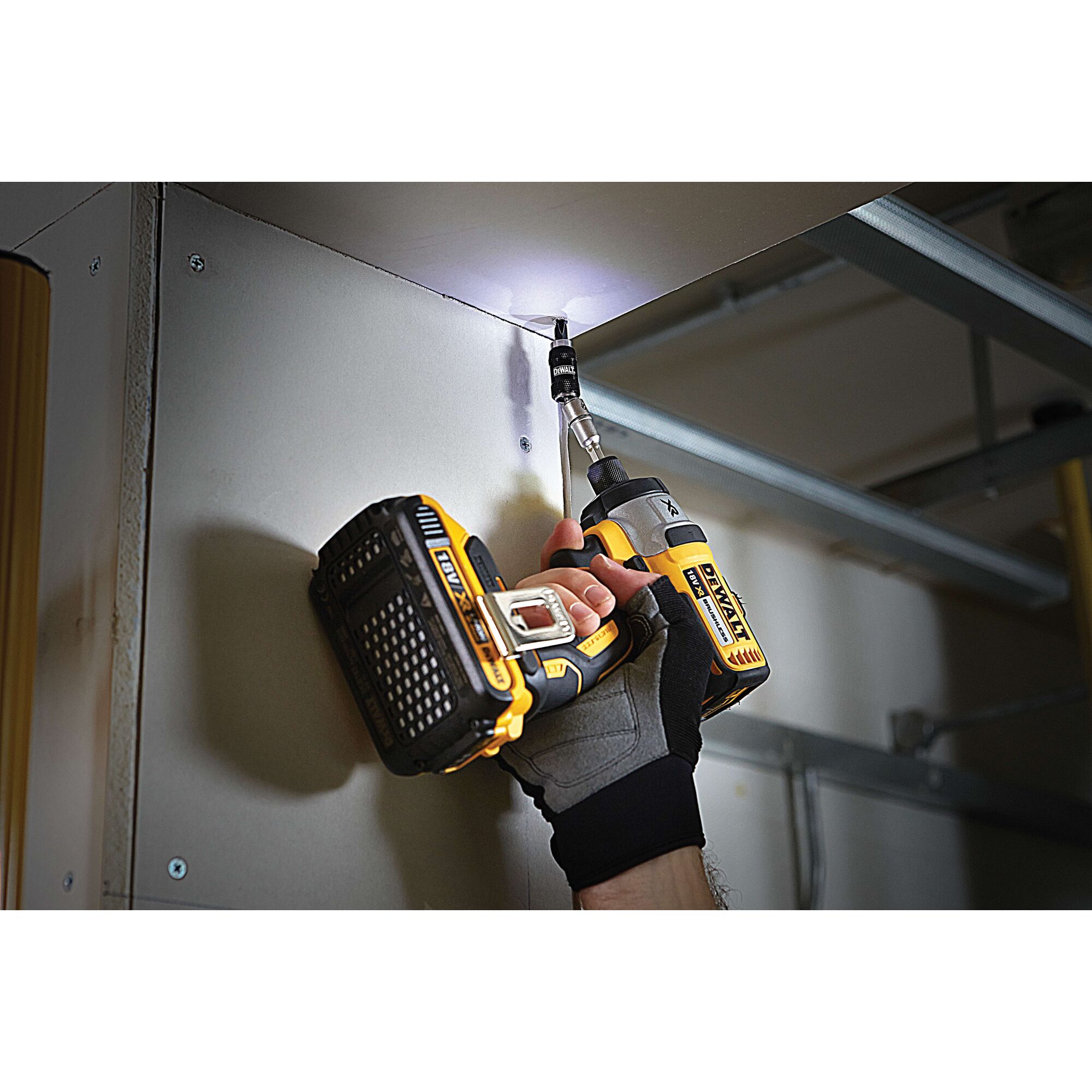 A person wearing work gloves uses a DEWALT cordless power drill to drive a screw into a ceiling corner, with a built-in LED light illuminating the work area.