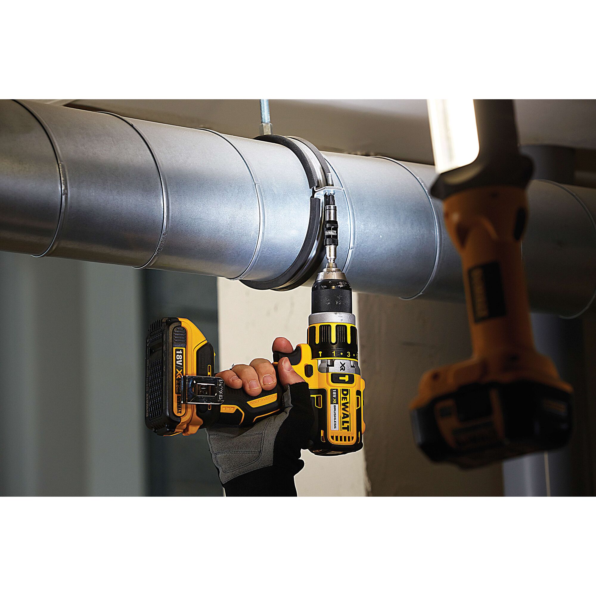 A DEWALT cordless drill is being used by a person wearing work gloves to fasten a clamp on a large metal duct pipe. Another DEWALT tool is partially visible in the foreground.