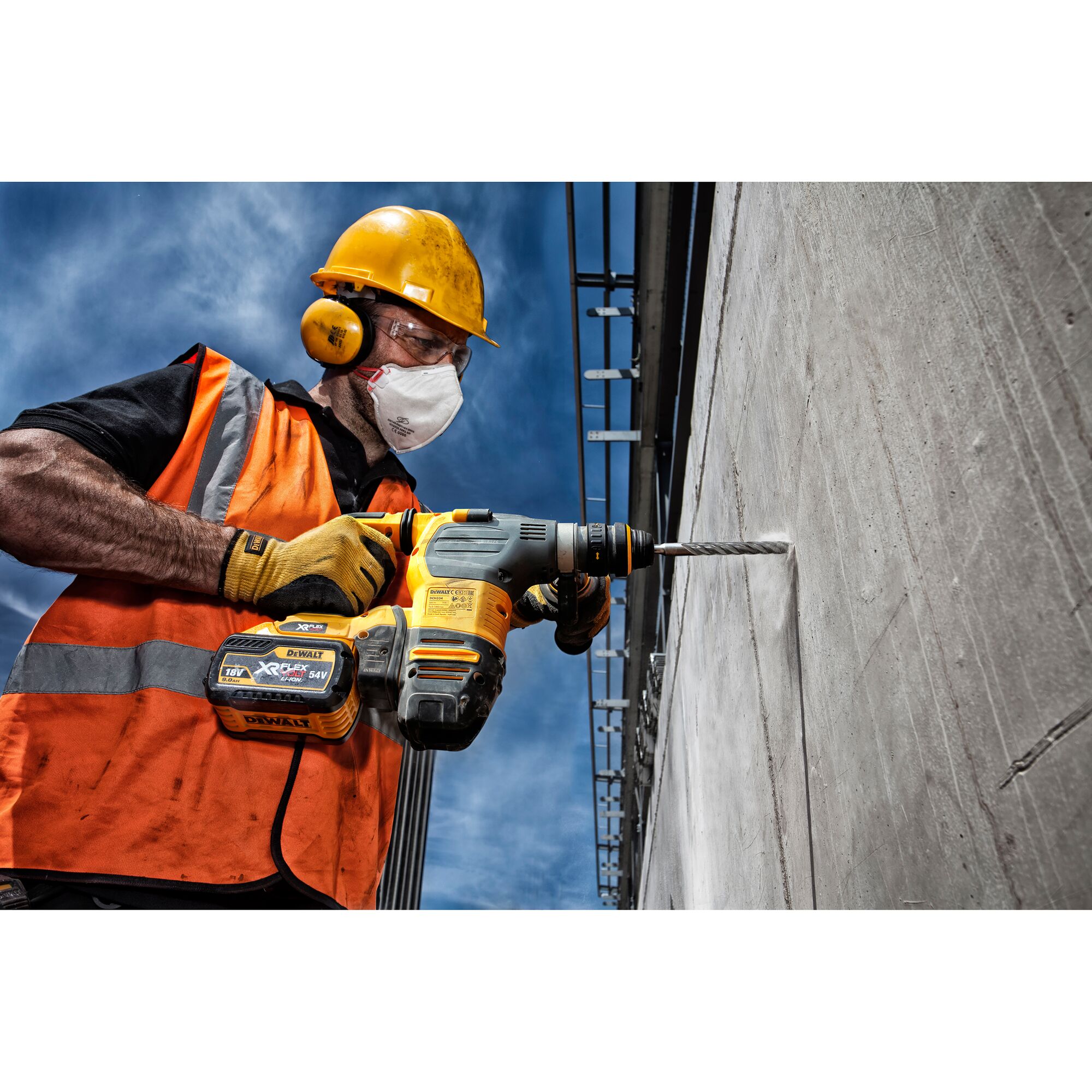 A construction worker wearing a yellow hard hat, orange safety vest, and yellow gloves is using a DEWALT cordless hammer drill to drill into a concrete wall at an outdoor construction site. The worker's face is not visible.