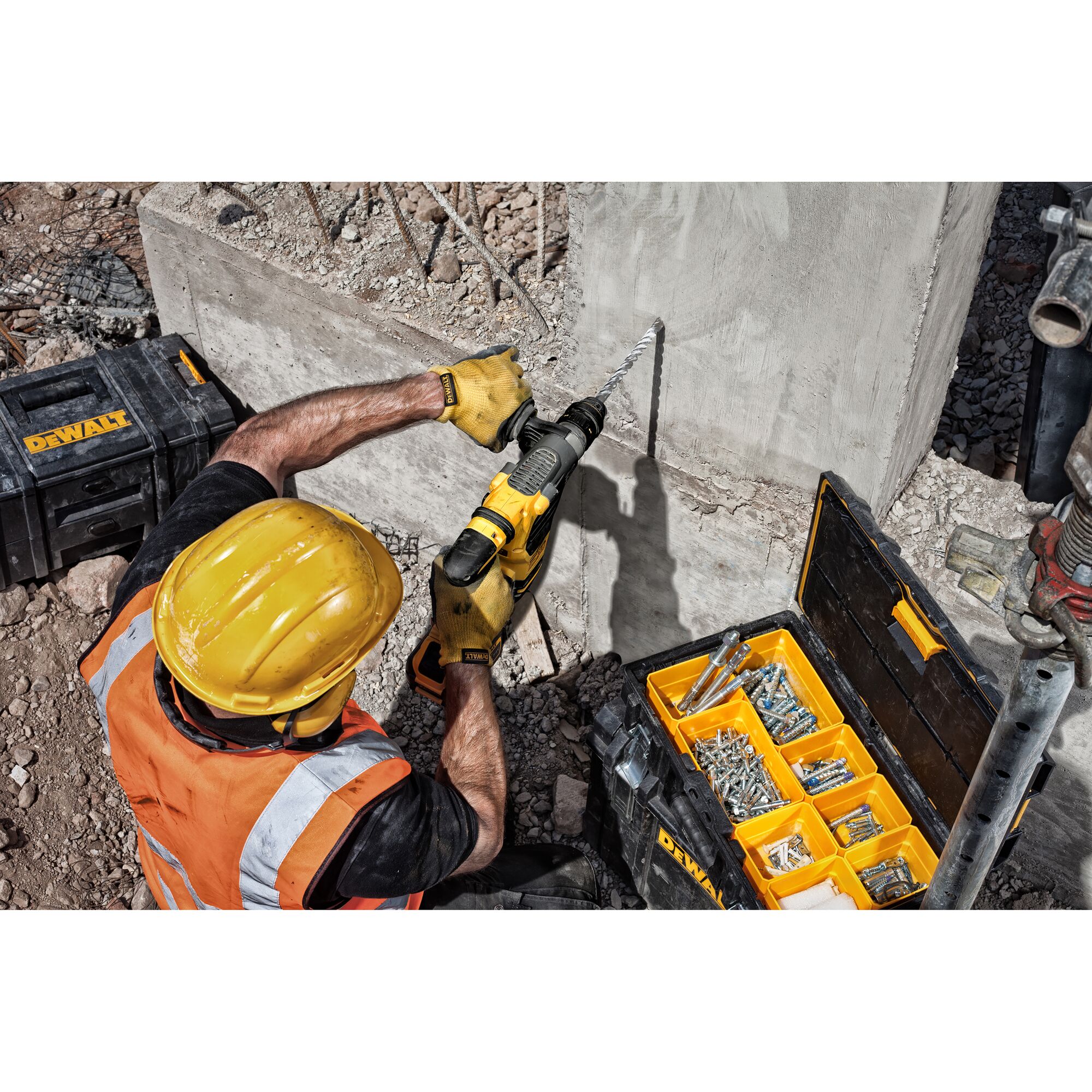 A construction worker wearing a yellow hard hat and orange safety vest uses a DEWALT hammer drill to drill into a concrete wall. An open DEWALT tool box with organized screws and anchors is on the ground nearby.
