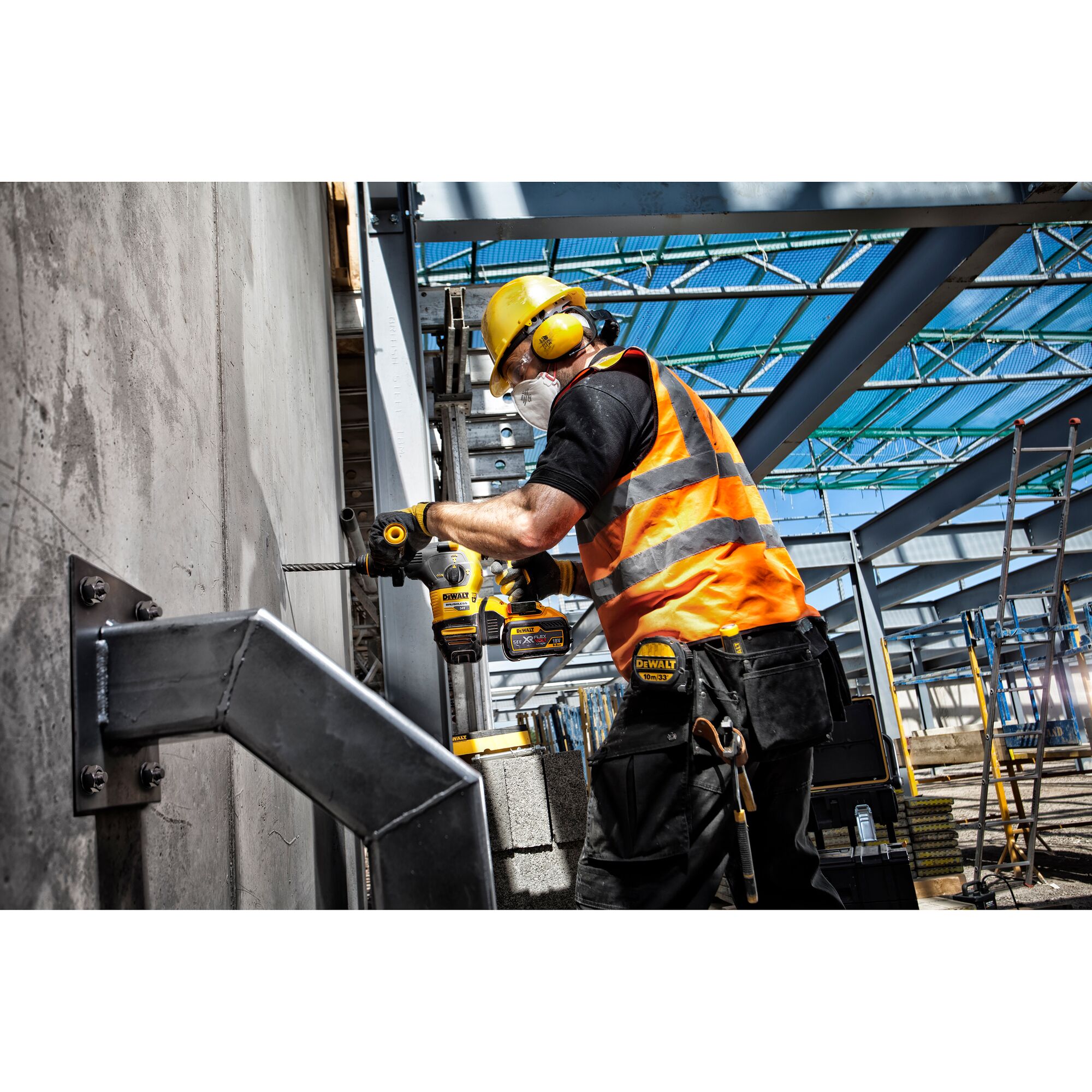 A construction worker wearing a yellow hard hat, orange reflective vest, ear protection, and face mask is using a DEWALT power drill to drill into a concrete wall on a construction site. Metal beams and construction materials are visible in the background.