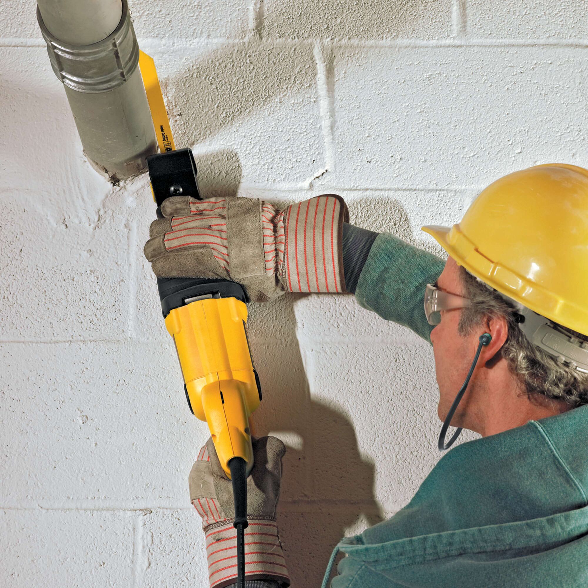 A person wearing safety gloves, goggles, and a yellow hard hat uses a yellow DEWALT reciprocating saw (model DW304PK) to cut through a metal pipe attached to a white brick wall.