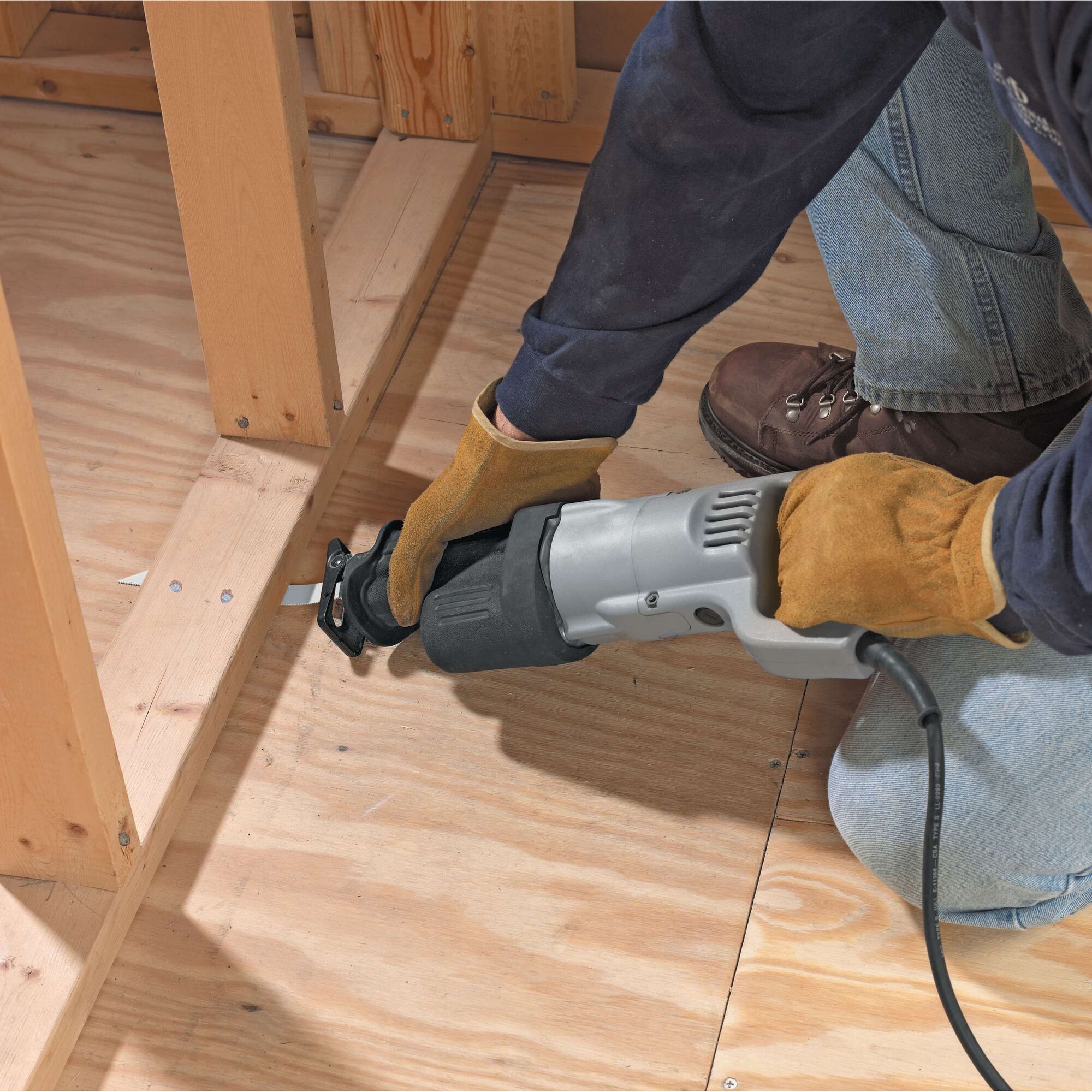 A person wearing gloves and work boots is using a corded reciprocating saw to cut a piece of wood on a plywood floor inside a construction frame.