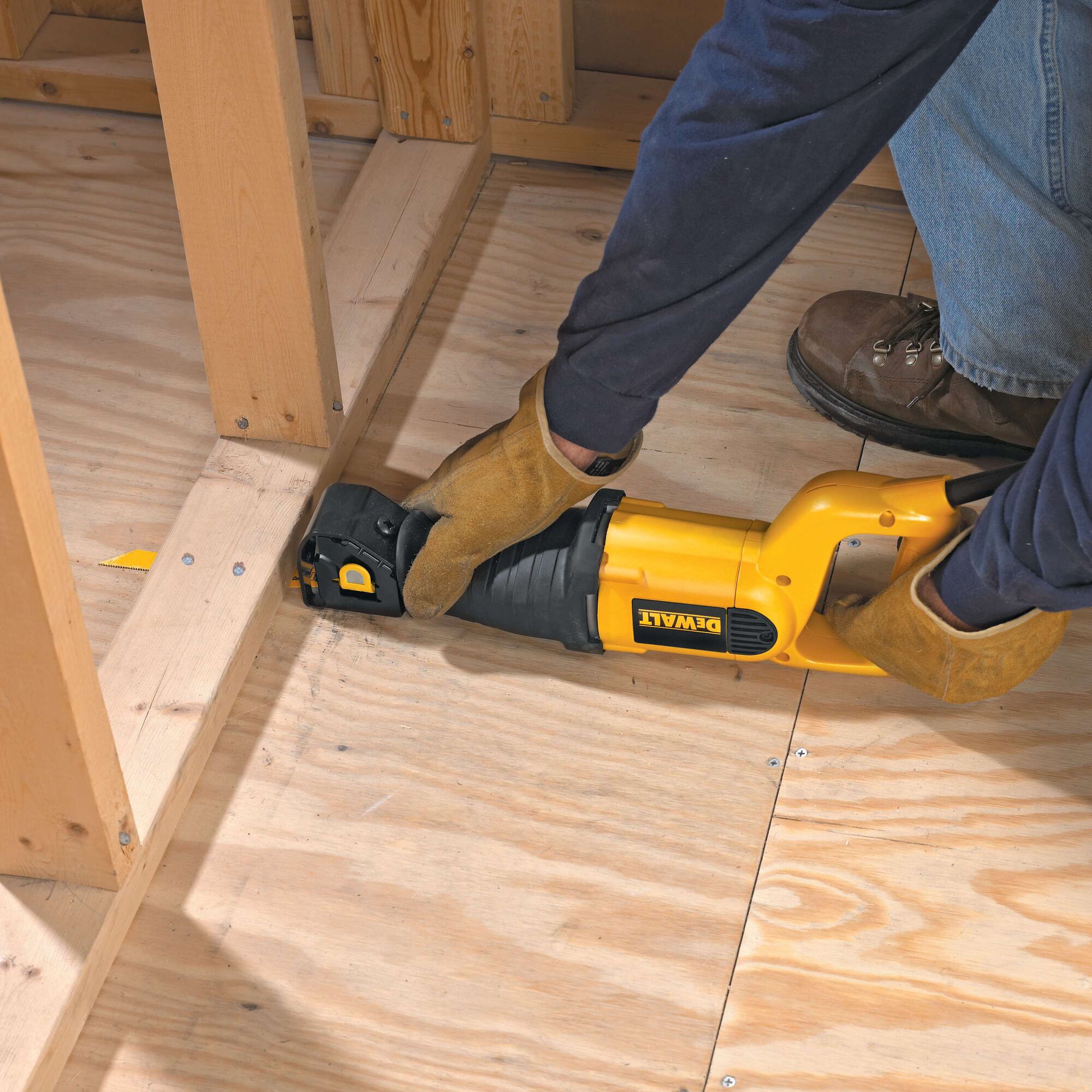 A person wearing gloves and work boots uses a yellow DEWALT reciprocating saw to cut plywood flooring near wooden framing.
