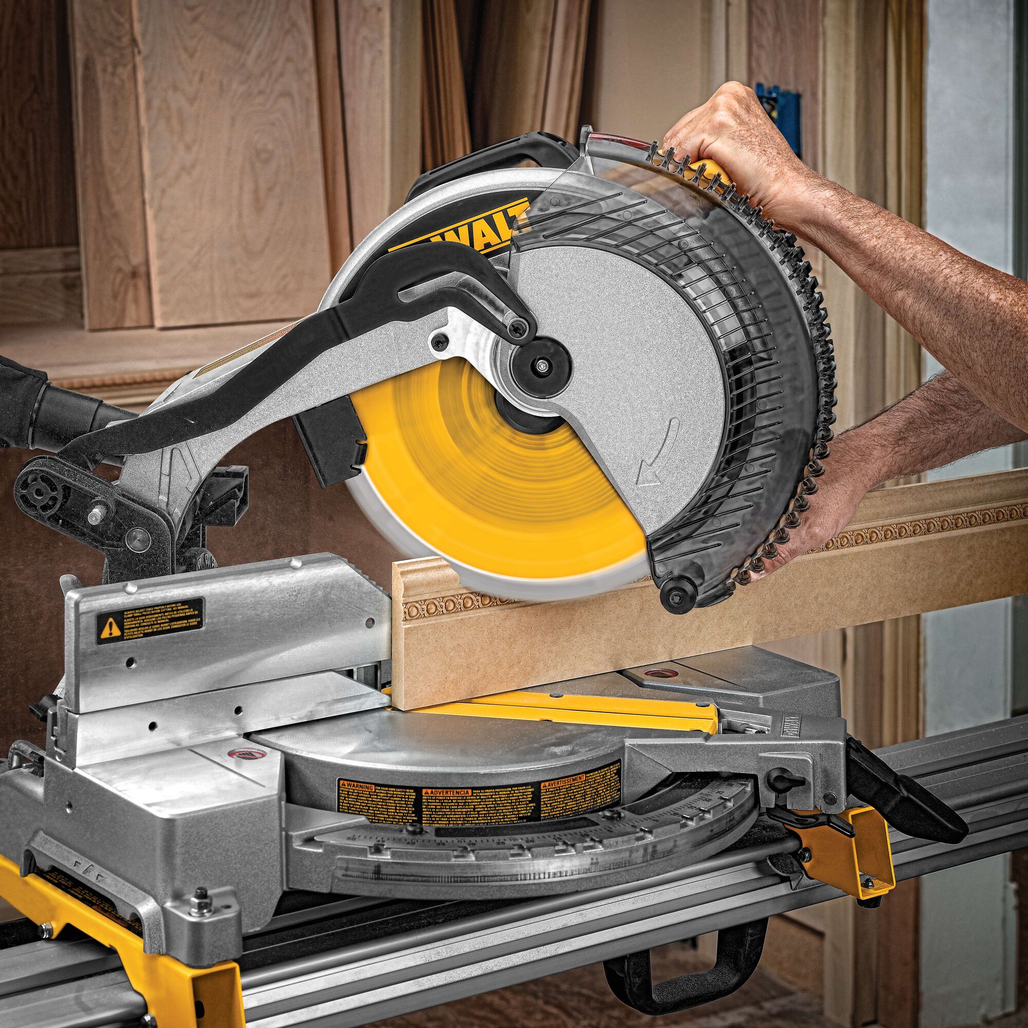 A DEWALT miter saw is cutting a piece of wood. The saw's circular blade is spinning, and a person's hand is operating the tool. The workspace is surrounded by wooden panels and boards.
