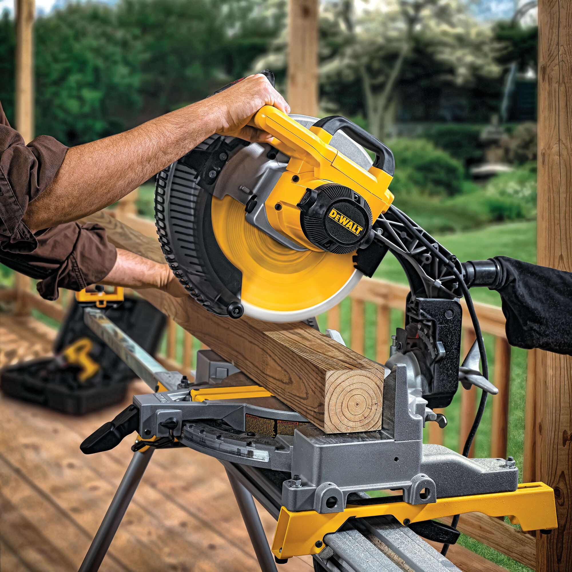 A person is using a DEWALT miter saw to cut a wooden beam on a deck outdoors. The saw is mounted on a stand and the DEWALT brand logo is visible on the saw. The setting includes natural greenery in the background.